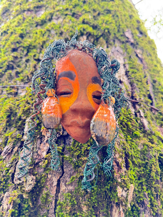 Adda's unique design against a mossy tree bark backdrop is sure to catch your eye. This 3” x 5” 6oz. mixed media raku fire mask, crafted with 15 feet of 24 and 16 gauge wire, adds a touch of elegance to your HerDew mask collection. With a copper nose ring, three-colored complexion, and a large Raku fired earring with two wooden beads, Adda is a must-have for any mask enthusiast.