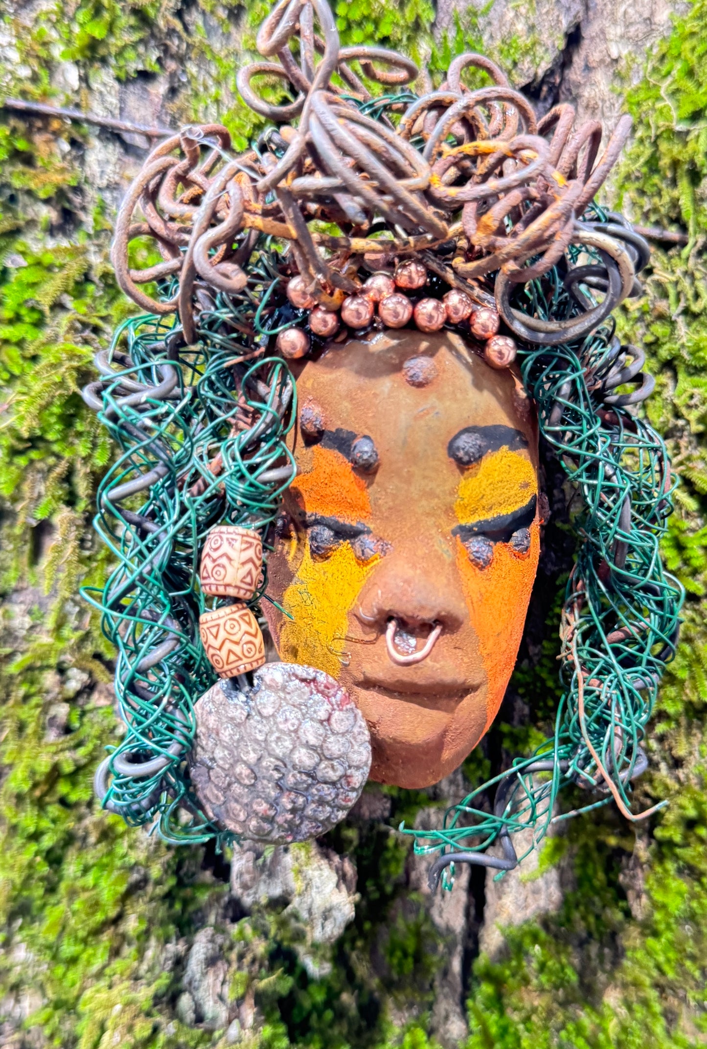 Adda's unique design against a mossy tree bark backdrop is sure to catch your eye. This 3” x 5” 6oz. mixed media raku fire mask, crafted with 15 feet of 24 and 16 gauge wire, adds a touch of elegance to your HerDew mask collection. With a copper nose ring, three-colored complexion, and a large Raku fired earring with two wooden beads, Adda is a must-have for any mask enthusiast.