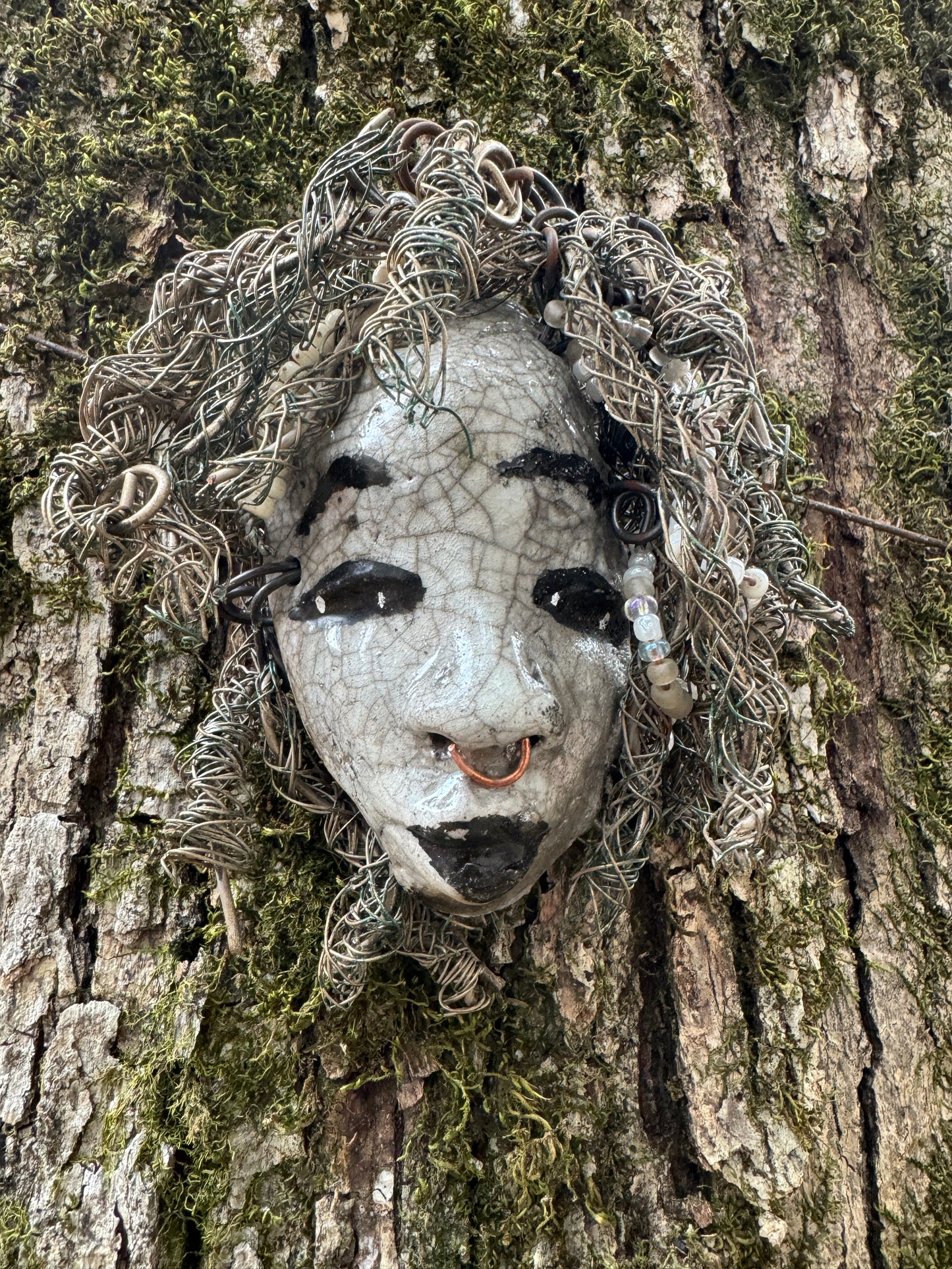 ami is a 4x 6” raku-fired mask featuring a pristine off-white color palette. Her intricately detailed hair is composed of nearly 15 feet of 16 gauge wire, delicately adorned with dainty beads. Set in a backdrop of nature, her portrait makes a wonderful addition to any outdoor art collector, or for decorating the home.