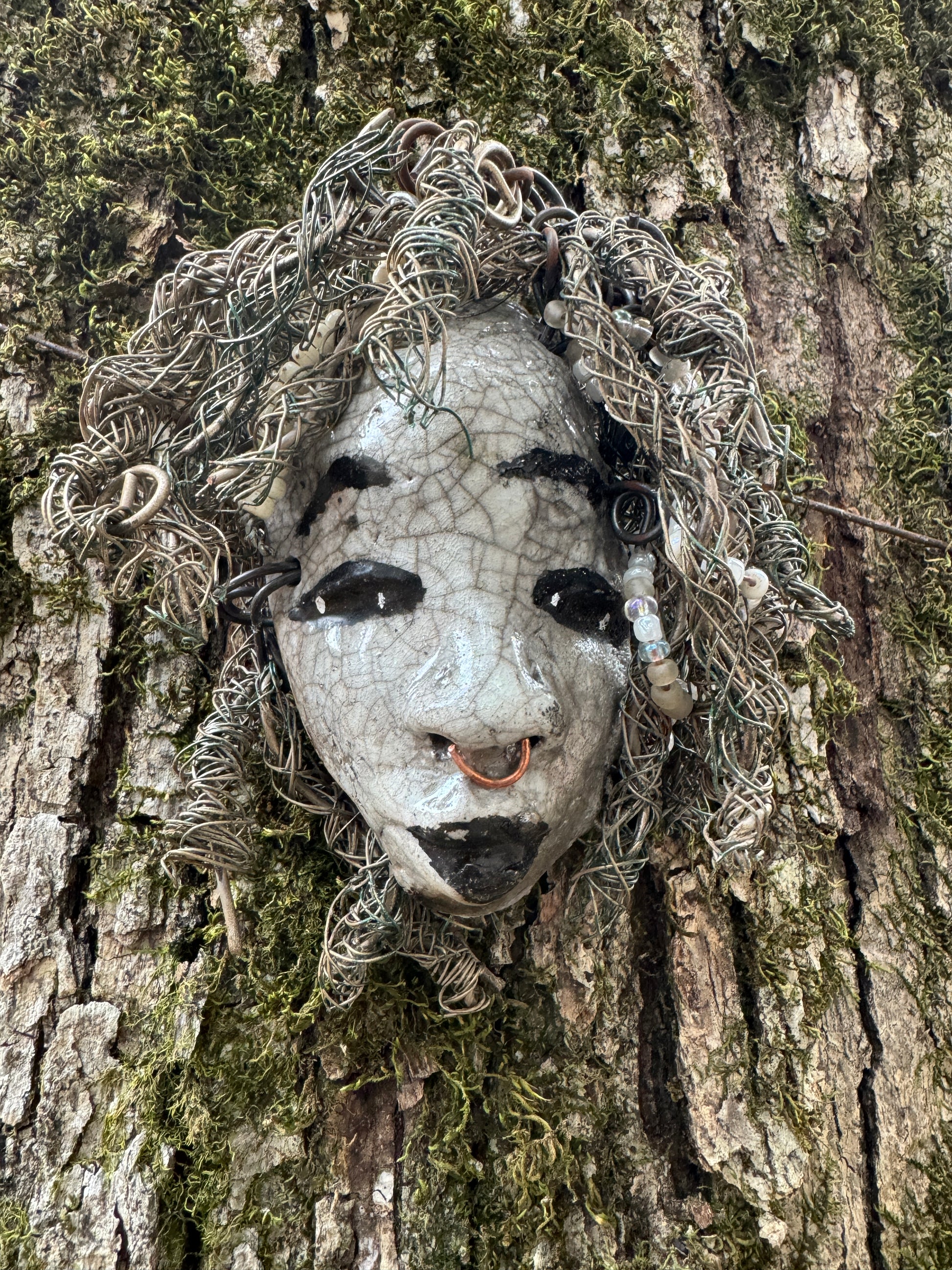 ami is a 4x 6” raku-fired mask featuring a pristine off-white color palette. Her intricately detailed hair is composed of nearly 15 feet of 16 gauge wire, delicately adorned with dainty beads. Set in a backdrop of nature, her portrait makes a wonderful addition to any outdoor art collector, or for decorating the home.