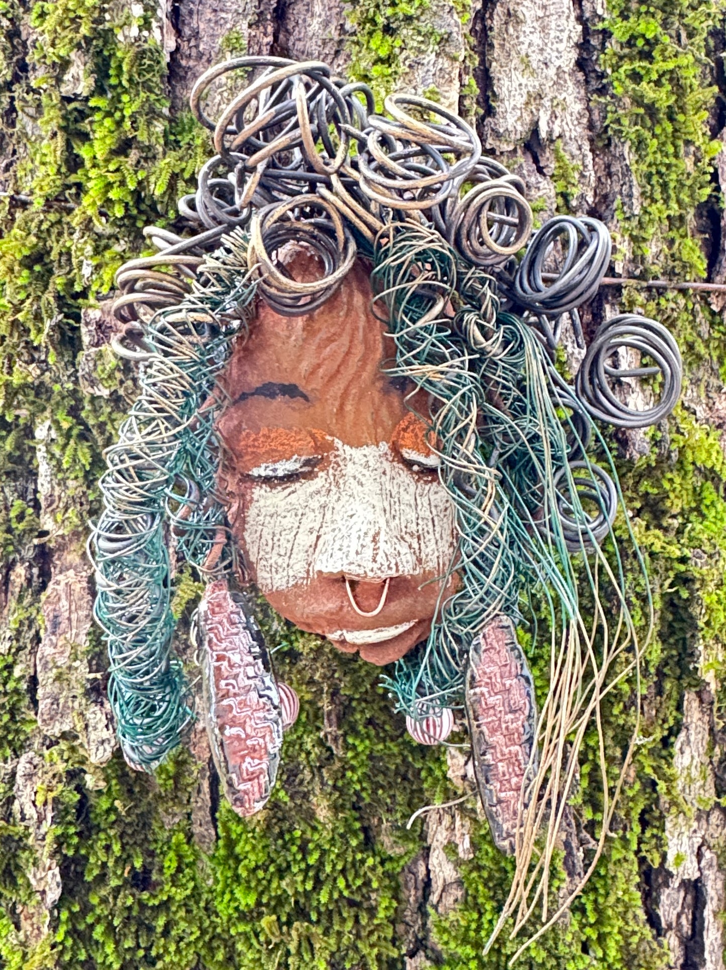 Baldwin Bo is a mixed media raku fire mask, measuring 4" x 6" and weighing 6oz. This unique piece features 25 feet of 24 and 16 gauge wire and a copper nose ring, giving it a three-colored complexion. The mask is also adorned with large Raku fired earrings and a wooden bead, and looks stunning when photographed against a mossy tree bark. Add Baldwin Bo to your collection of HerDew masks for a beautiful and eye-catching addition.