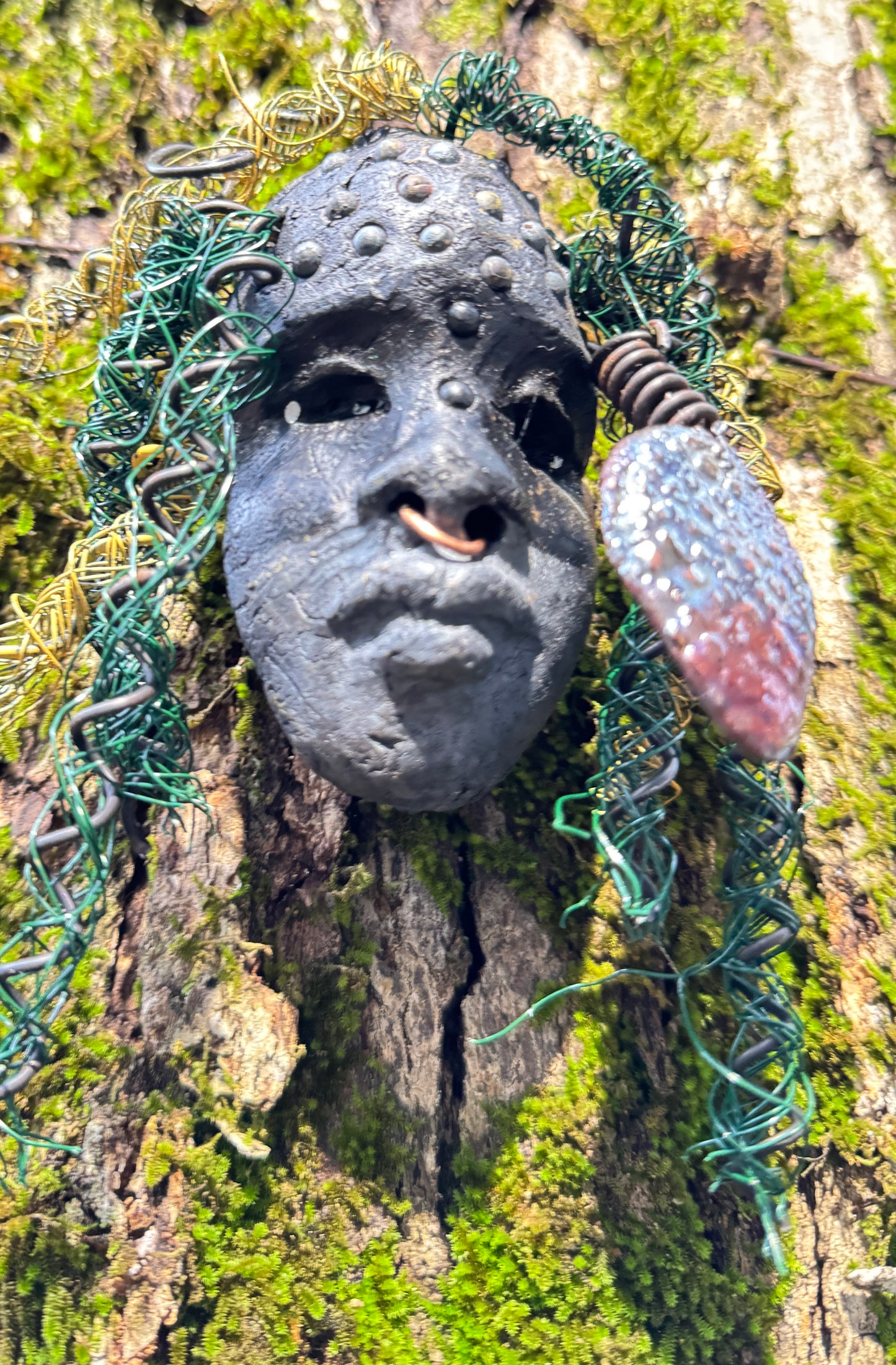 Enhance your HerDew mask collection with Kao! This mixed media raku fire mask is 4” x 6” and 6oz., featuring 25 feet of 24 and 16 gauge wire and a copper nose ring. Its exotic appeal is captured in a photo on tree bark, adding a unique touch to your collection. The mask's Smokey black complexion and Raku fired earring with a wooden bead make it a truly stunning piece..