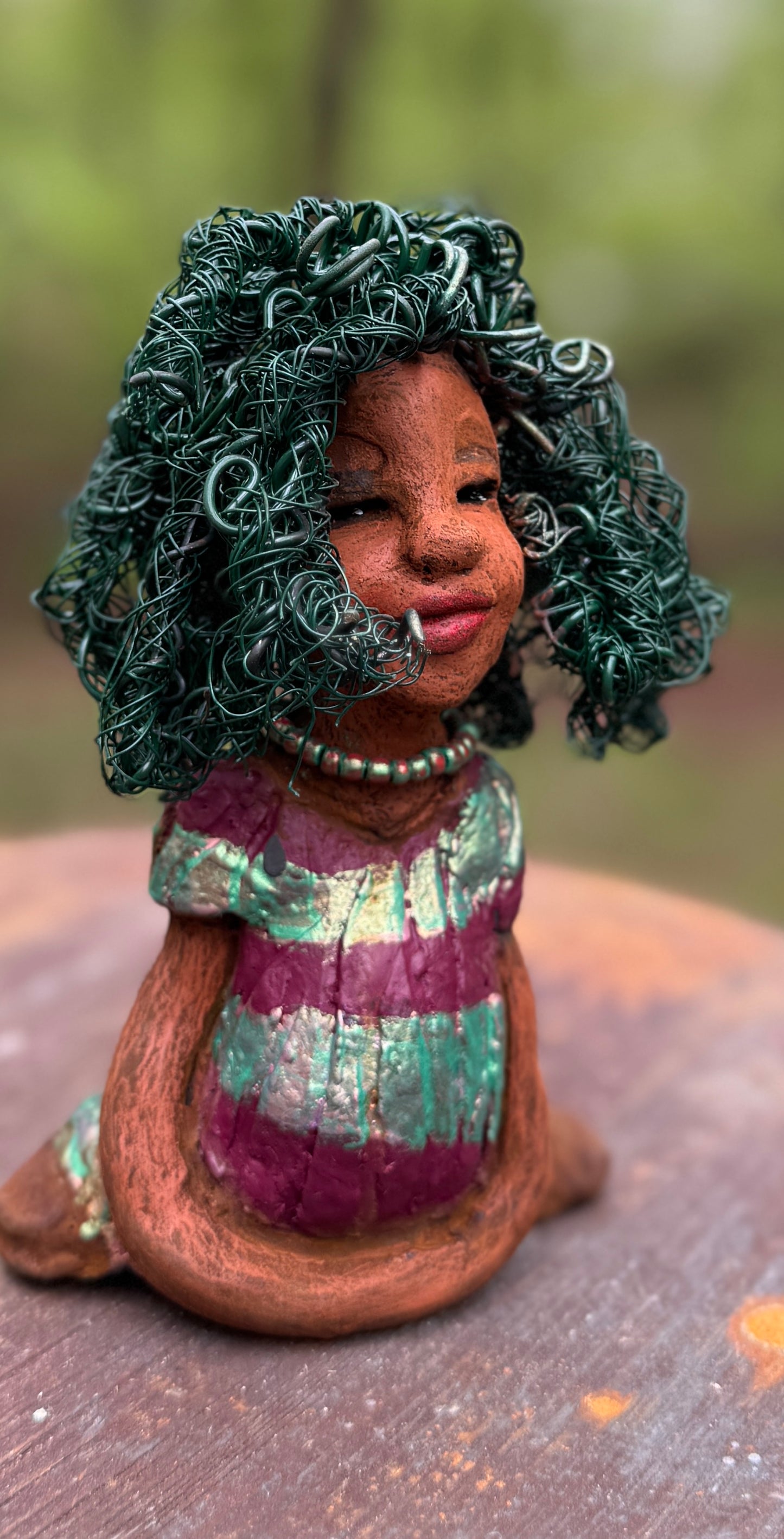 Coco is a 7.5" x 4" x 3" figurine that weighs 1 lb and wears a stunning red and green dress with stripes. Her wire hair is intricately coiled and adds to her regal appearance. Poised in a yoga pose, Coco's expression exudes curiosity, making her a perfect addition to the Herdew Collection.