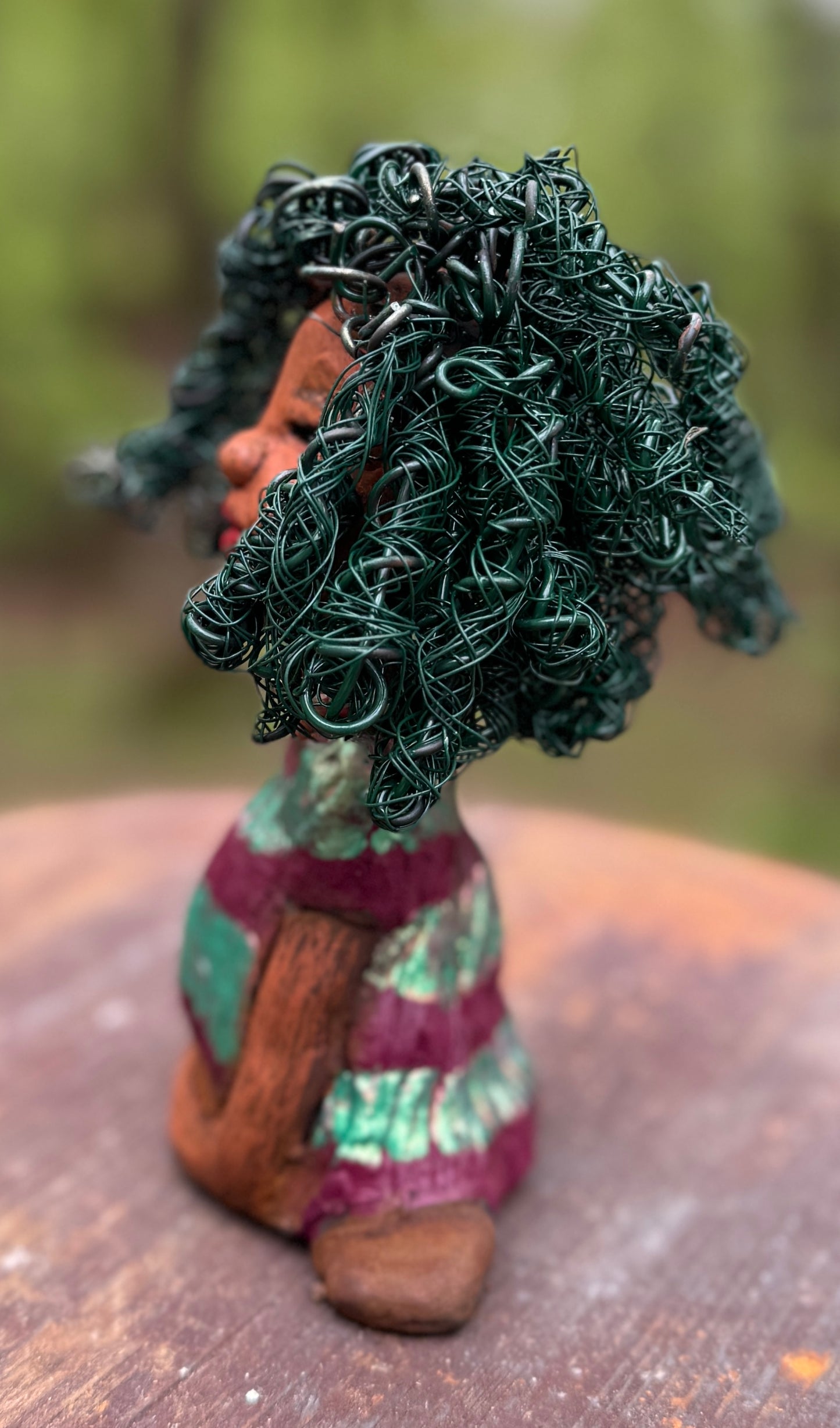 Coco is a 7.5" x 4" x 3" figurine that weighs 1 lb and wears a stunning red and green dress with stripes. Her wire hair is intricately coiled and adds to her regal appearance. Poised in a yoga pose, Coco's expression exudes curiosity, making her a perfect addition to the Herdew Collection.