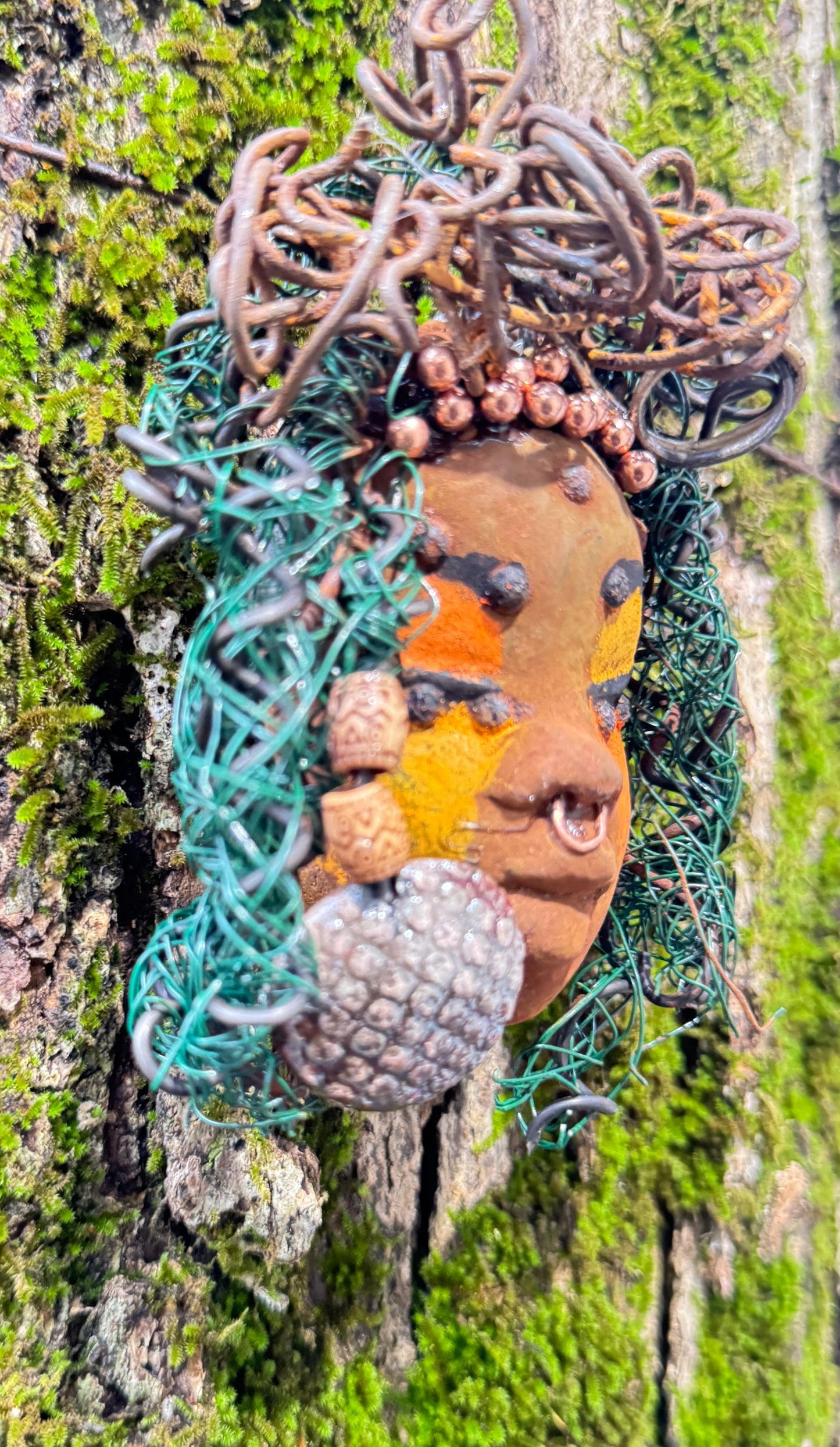 Adda's unique design against a mossy tree bark backdrop is sure to catch your eye. This 3” x 5” 6oz. mixed media raku fire mask, crafted with 15 feet of 24 and 16 gauge wire, adds a touch of elegance to your HerDew mask collection. With a copper nose ring, three-colored complexion, and a large Raku fired earring with two wooden beads, Adda is a must-have for any mask enthusiast.