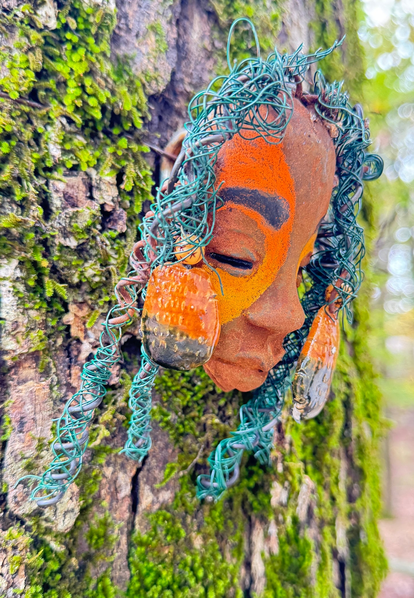 Adda's unique design against a mossy tree bark backdrop is sure to catch your eye. This 3” x 5” 6oz. mixed media raku fire mask, crafted with 15 feet of 24 and 16 gauge wire, adds a touch of elegance to your HerDew mask collection. With a copper nose ring, three-colored complexion, and a large Raku fired earring with two wooden beads, Adda is a must-have for any mask enthusiast.