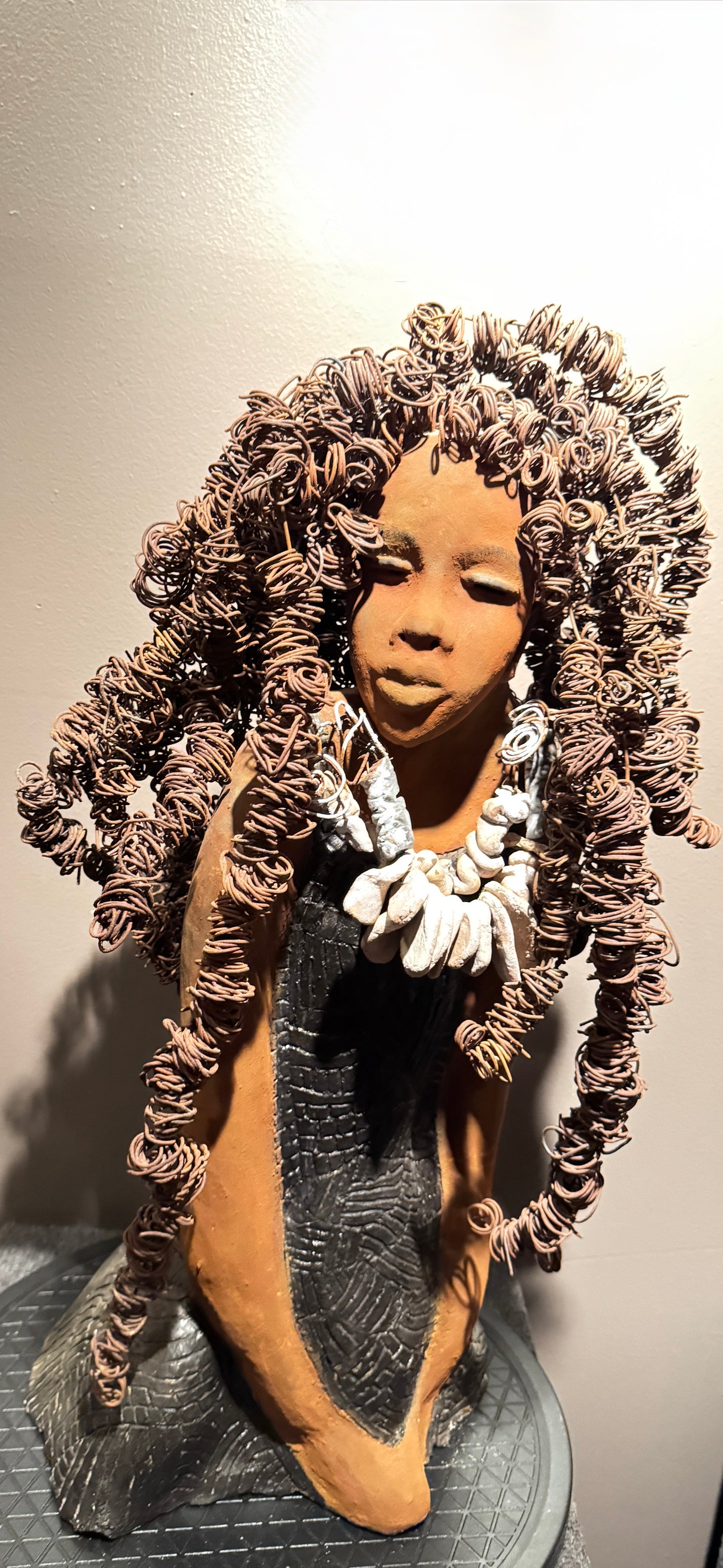 This evocative sculpture by Lester Jones celebrates the beauty, strength, and cultural heritage of African-descended women through form, texture, and symbolism. The figure’s elaborate crown of coiled wire hair evokes vitality, creativity, and a sense of reverence. Her closed eyes and serene expression suggest introspection, grace, or spiritual grounding. Adorned with a necklace clay shell-like forms and dressed in a patterned robe, the sculpture bridges ancestral tradition and modern artistry.
Lester J