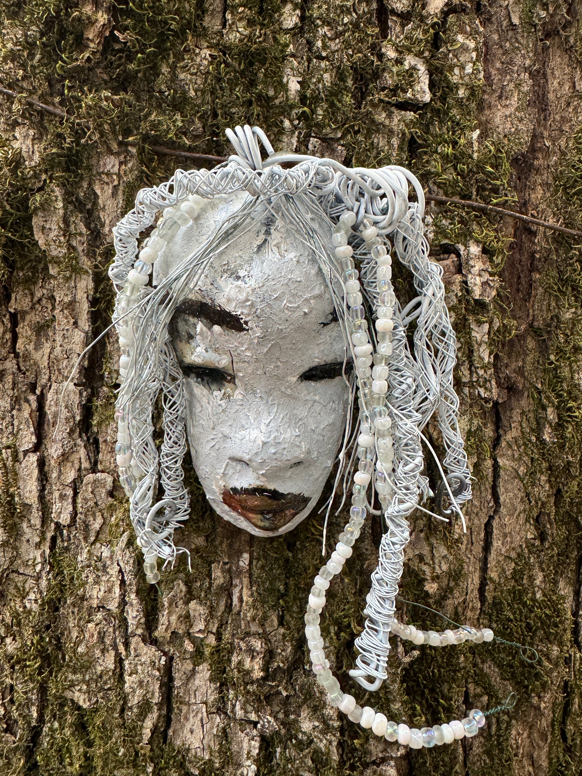 Olivia is a 4 x 7” 5.5 oz raku-fired mask featuring an off-white complexion. 16 gauge wire measuring over 15 feet is used as her hair, accessorized with small decorative beads. Her portrait was taken in an outdoor setting featuring a tree, making her an ideal gift for those who enjoy outdoor art or want to adorn the inside of their home.