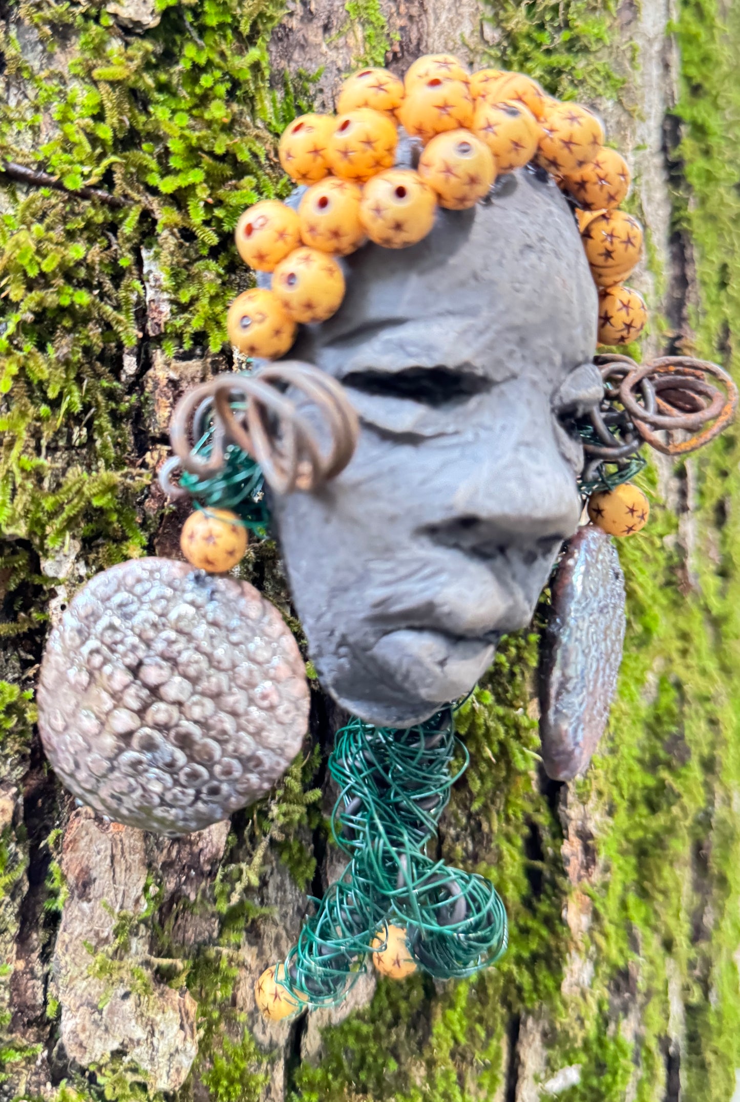Introducing Cali, a 3” x 5” 5oz. mixed media raku fire mask adorned with a copper nose ring, three-colored complexion, and two large Raku fired earrings with a wooden bead. With 15 feet of 24 and 16 gauge wire, this piece is a stunning addition to your HerDew mask collection. See the beautiful Cali against a tree bark background.