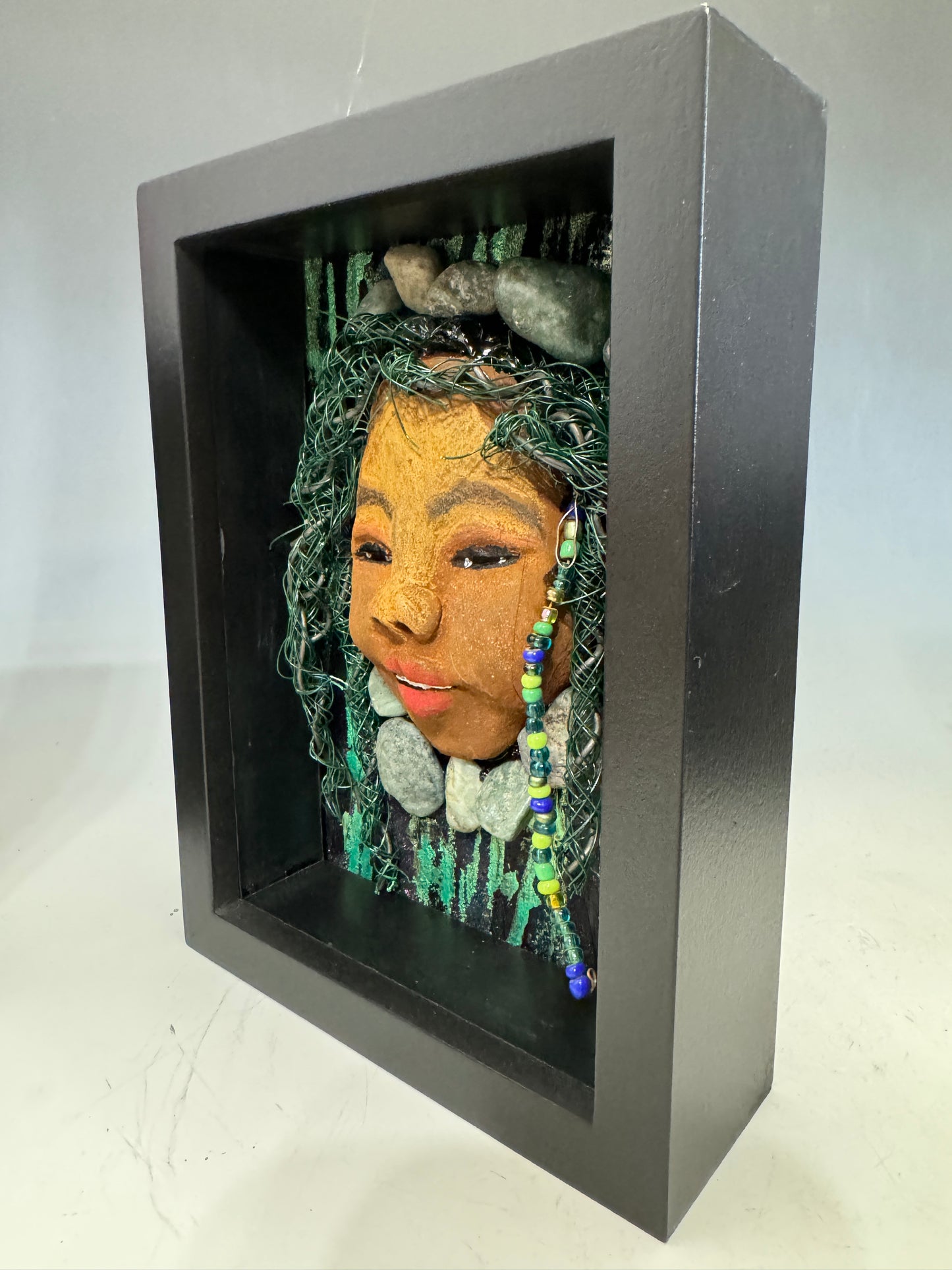Introducing Allie, a mixed media sculpture raku fired mask. Her features include a two-tone honey brown complexion, Ruby red lips, and hair intricately intertwined with 16 and 24 gauge wire. She is mounted on a hand-painted canvas, accented by landscaped background, and enclosed in a 5“ x 7“ black shadowbox.