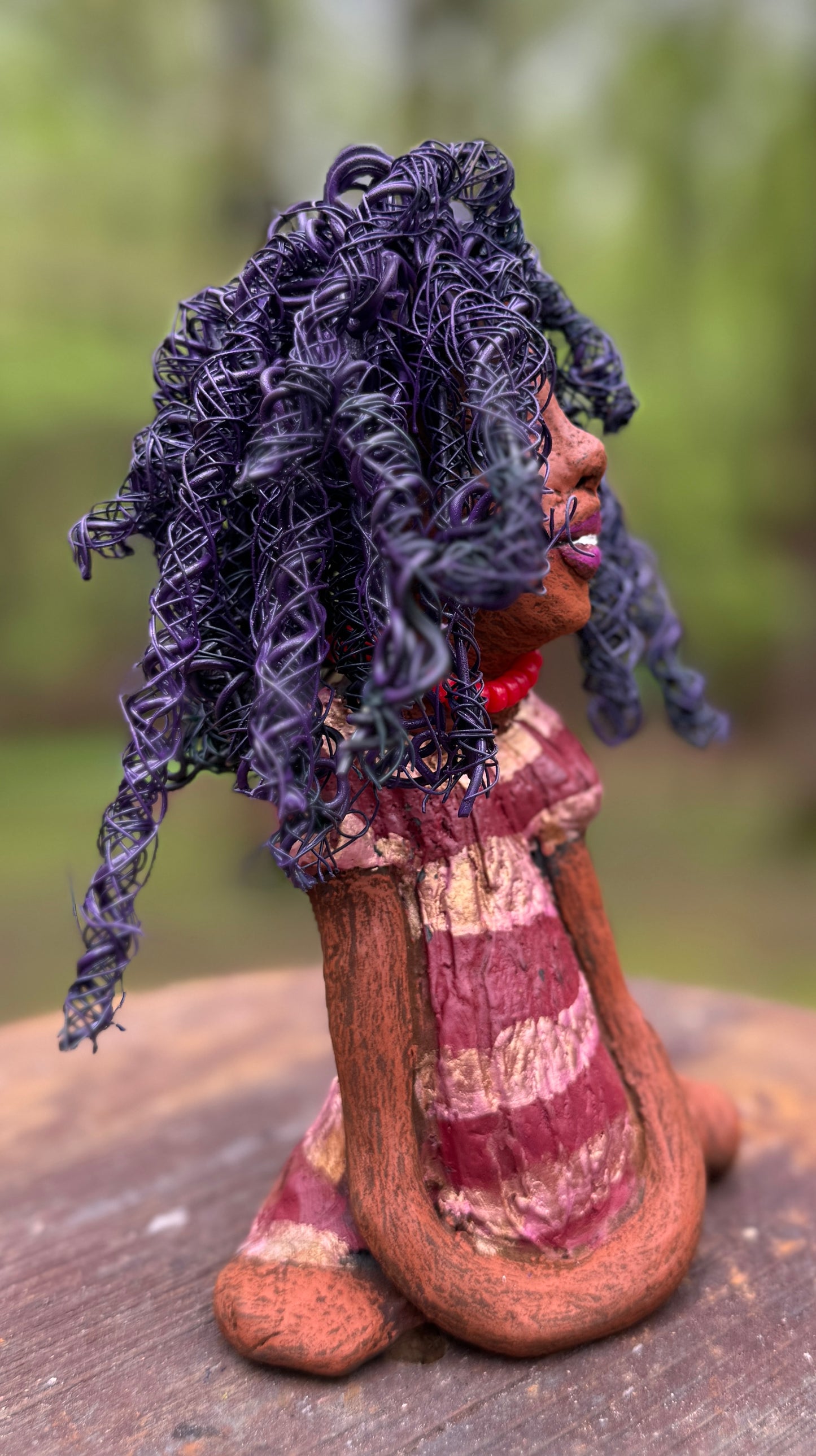 Introducing LaDonya, who measures 8" x 4. 5" x 5.5" and weighs 1.27 lbs. Her beauty is adorned with a lovely honey brown complexion, soft dark red lips, and long twisted purple locs that cascade down to her waist. Her metallic striped copper gold glazed dress adds a luxurious touch. With over 75 feet of 16 and 24 gauge wire for hair, it took over 6 hours to perfect her twisted hairstyle. Her hopeful expression and captivating eyes make her the perfect addition to any home. Plus, enjoy free shipping with our
