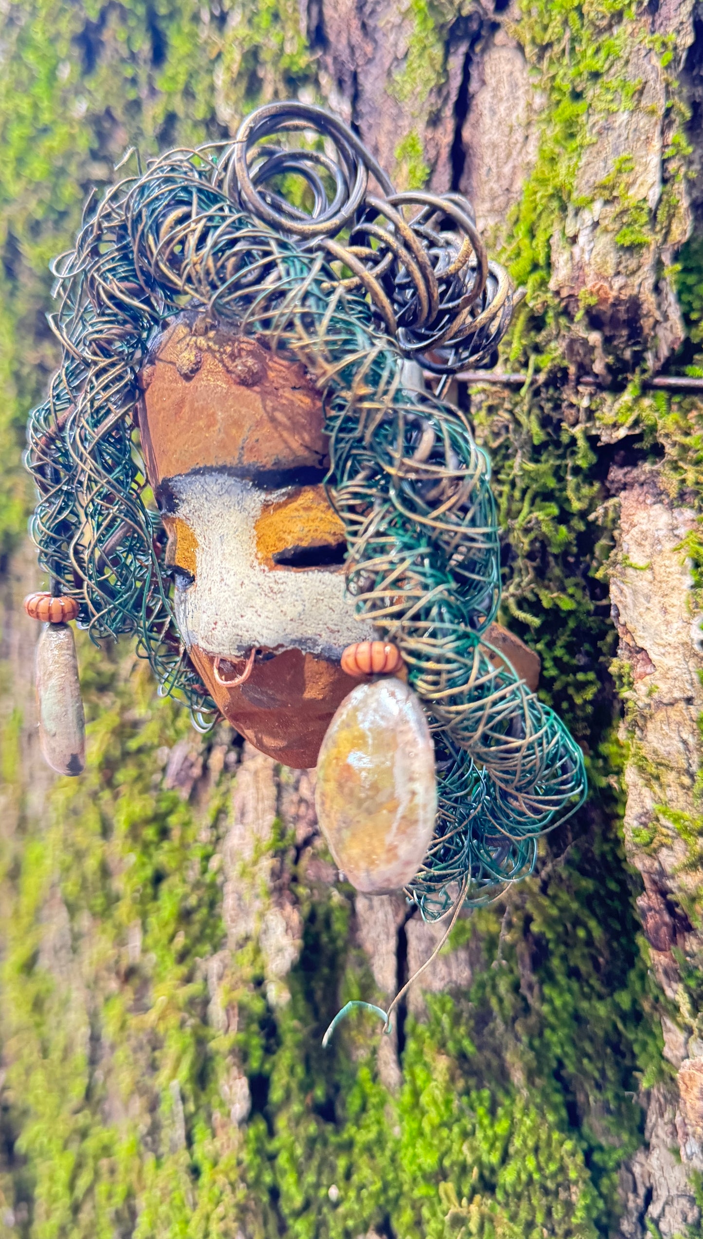 Alison is a 3” x 5” 5oz. mixed media raku fire mask with 15 feet of 24 and 16 gauge wire. Featuring a copper nose ring and three-colored complexion, this piece has two large Raku fired earrings with a wooden bead. Alison looks stunning with a mossy tree background and would make a lovely addition to your HerDew mask collection.