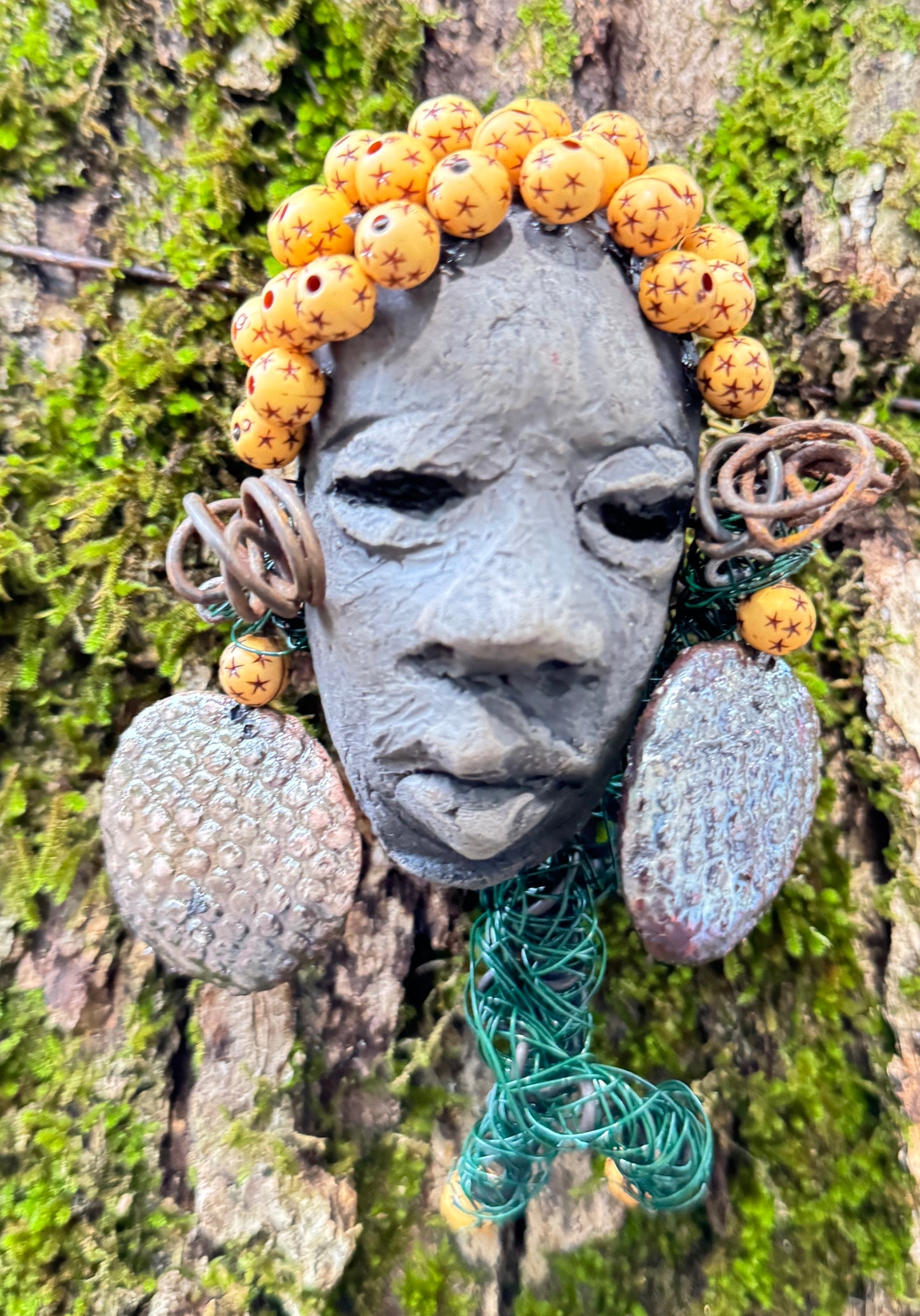 Introducing Cali, a 3” x 5” 5oz. mixed media raku fire mask adorned with a copper nose ring, three-colored complexion, and two large Raku fired earrings with a wooden bead. With 15 feet of 24 and 16 gauge wire, this piece is a stunning addition to your HerDew mask collection. See the beautiful Cali against a tree bark background.