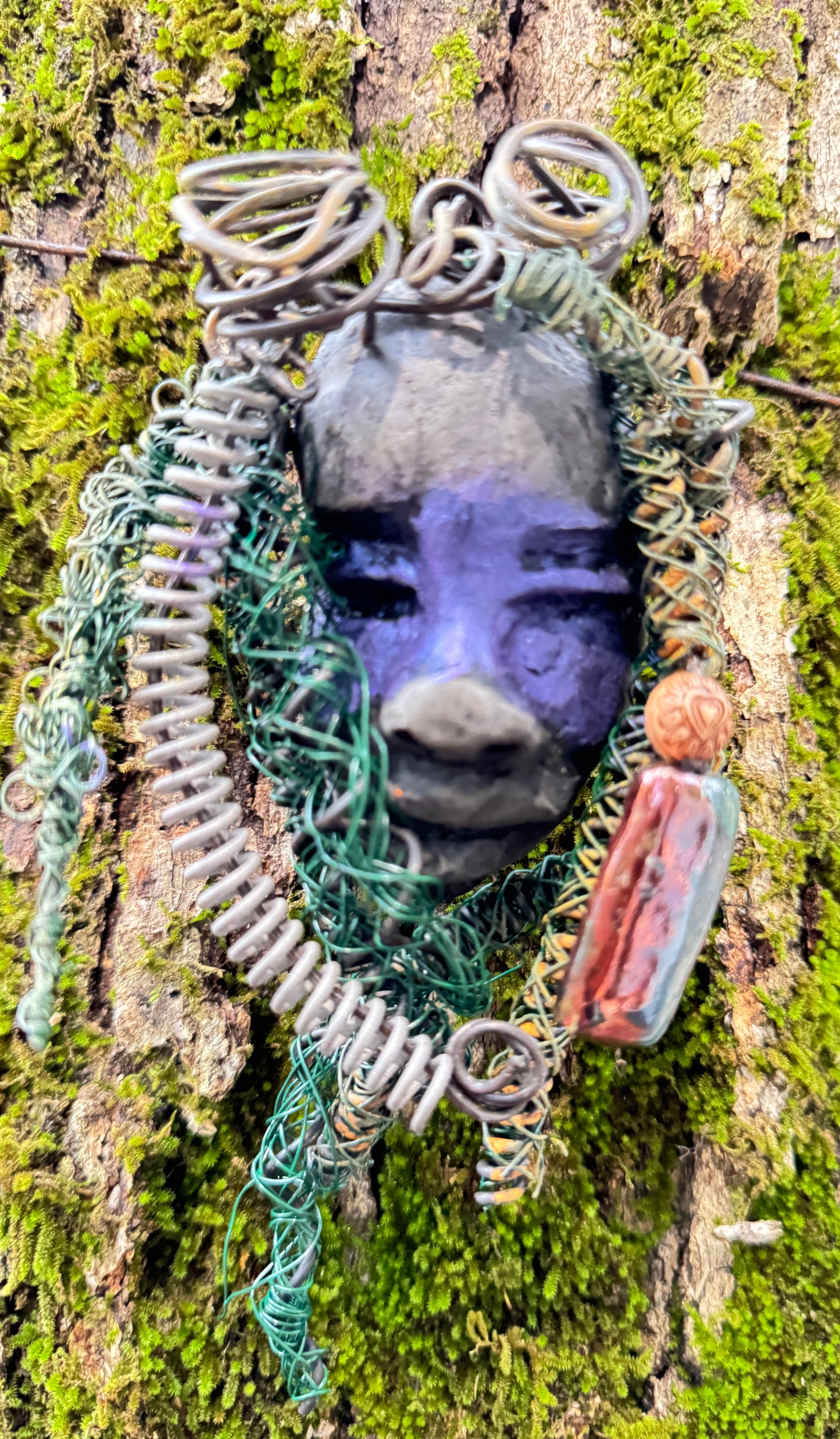 Rayda is a 4” x 6” 5oz. mixed media raku fire mask that includes 15 feet of 24 and 16 gauge wire. It has a Smokey black two tone complexion and a large metallic Raku fired earring with a wooden bead. Its earthy appeal is enhanced when displayed with natural tree bark, making it a lovely addition to your HerDew mask collection.