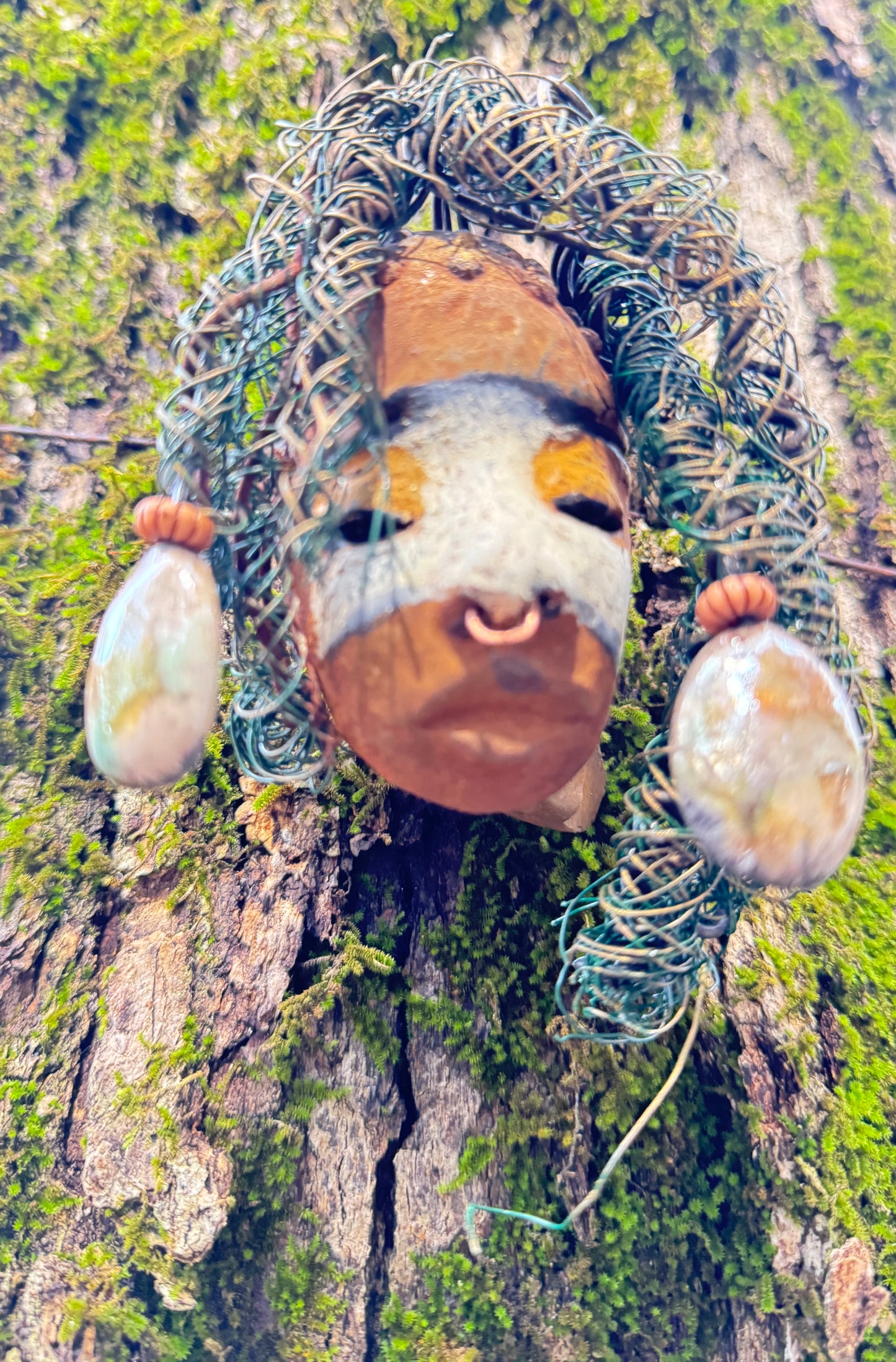 Alison is a 3” x 5” 5oz. mixed media raku fire mask with 15 feet of 24 and 16 gauge wire. Featuring a copper nose ring and three-colored complexion, this piece has two large Raku fired earrings with a wooden bead. Alison looks stunning with a mossy tree background and would make a lovely addition to your HerDew mask collection.