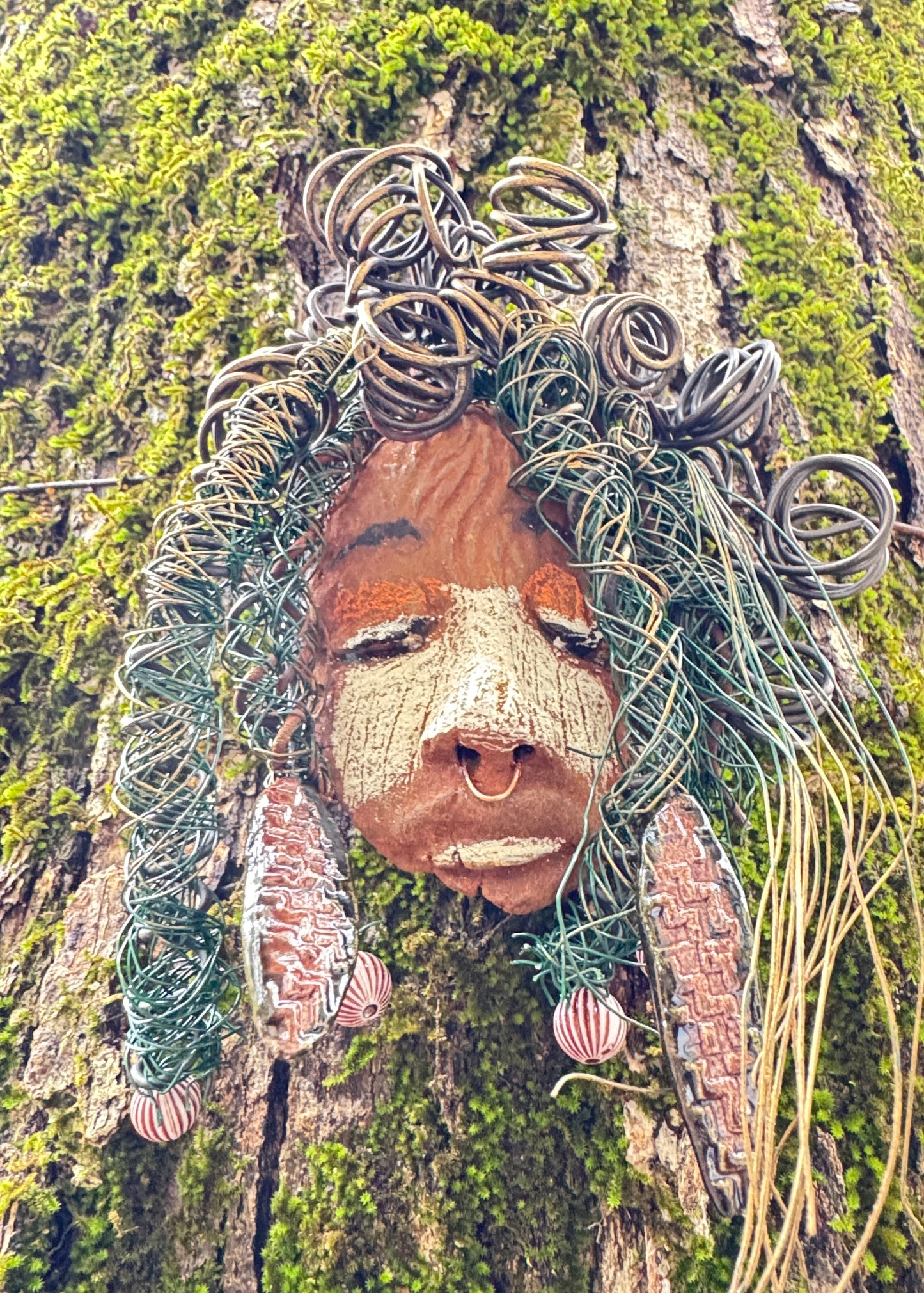 Baldwin Bo is a mixed media raku fire mask, measuring 4" x 6" and weighing 6oz. This unique piece features 25 feet of 24 and 16 gauge wire and a copper nose ring, giving it a three-colored complexion. The mask is also adorned with large Raku fired earrings and a wooden bead, and looks stunning when photographed against a mossy tree bark. Add Baldwin Bo to your collection of HerDew masks for a beautiful and eye-catching addition.