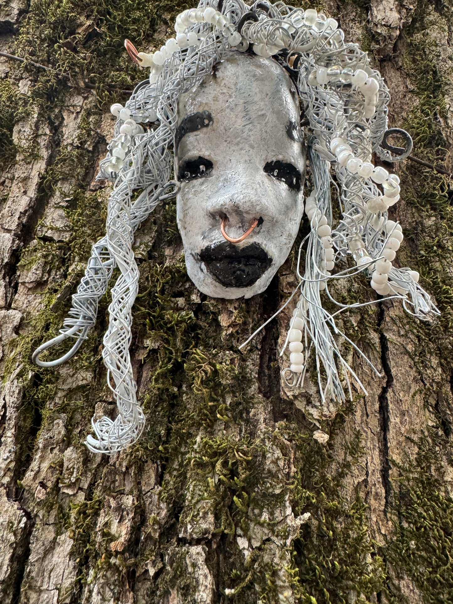 Juanita is a classic 4" x 4" 4.2 oz. Raku-fired mask designed with a white-hued complexion. Her hair is a long 15+ feet of 16-gauge wire, adorned with delicate beads. The portrait of her is set outdoors amongst a tree, making her an ideal present for art and nature admirers, as well as a perfect addition to any outdoor décor.