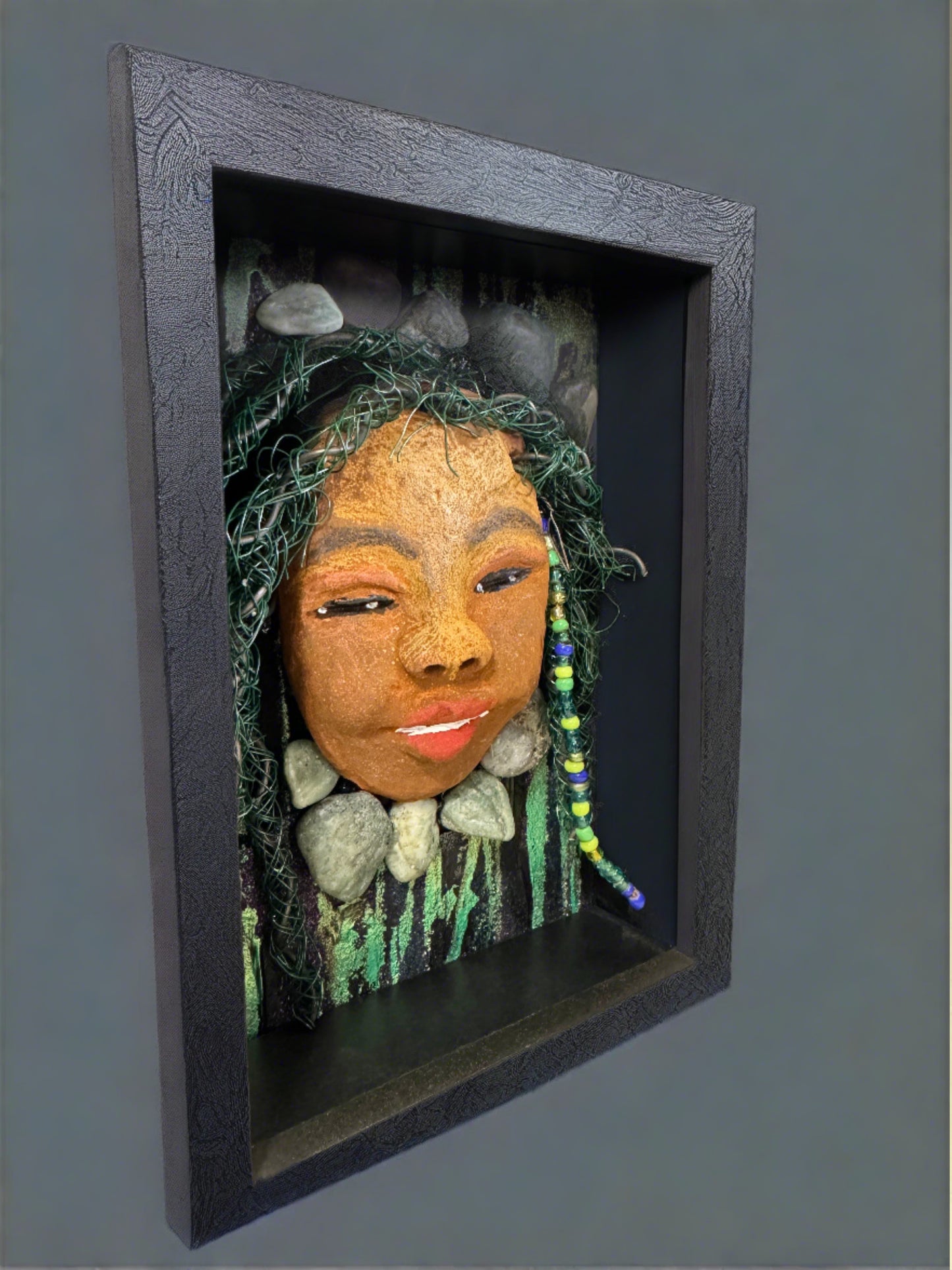 Introducing Allie, a mixed media sculpture raku fired mask. Her features include a two-tone honey brown complexion, Ruby red lips, and hair intricately intertwined with 16 and 24 gauge wire. She is mounted on a hand-painted canvas, accented by landscaped background, and enclosed in a 5“ x 7“ black shadowbox.