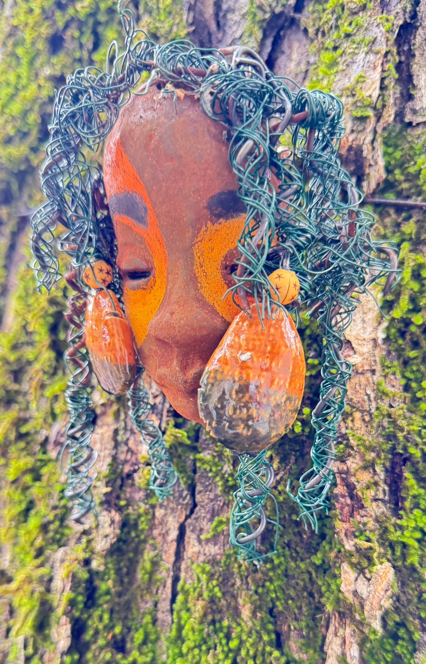 Adda's unique design against a mossy tree bark backdrop is sure to catch your eye. This 3” x 5” 6oz. mixed media raku fire mask, crafted with 15 feet of 24 and 16 gauge wire, adds a touch of elegance to your HerDew mask collection. With a copper nose ring, three-colored complexion, and a large Raku fired earring with two wooden beads, Adda is a must-have for any mask enthusiast.