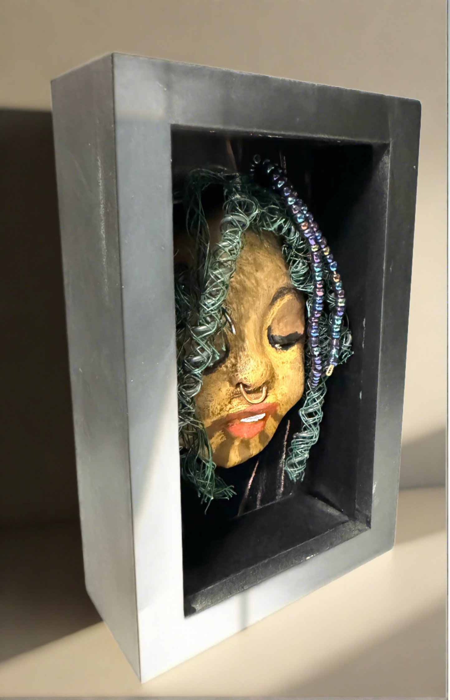 Introducing Baraka: An alluring mixed media sculpture created using the raku firing method. Impeccably designed attributes include a distinctive dual-toned honey brown complexion, delicate Ruby red lips, and intricately woven hair using 16 and 24 gauge wire. Mounted on a hand-painted canvas with a stunning landscape backdrop, this piece is elegantly showcased in a 5" x 7" black shadowbox, protected by a glass cover.
