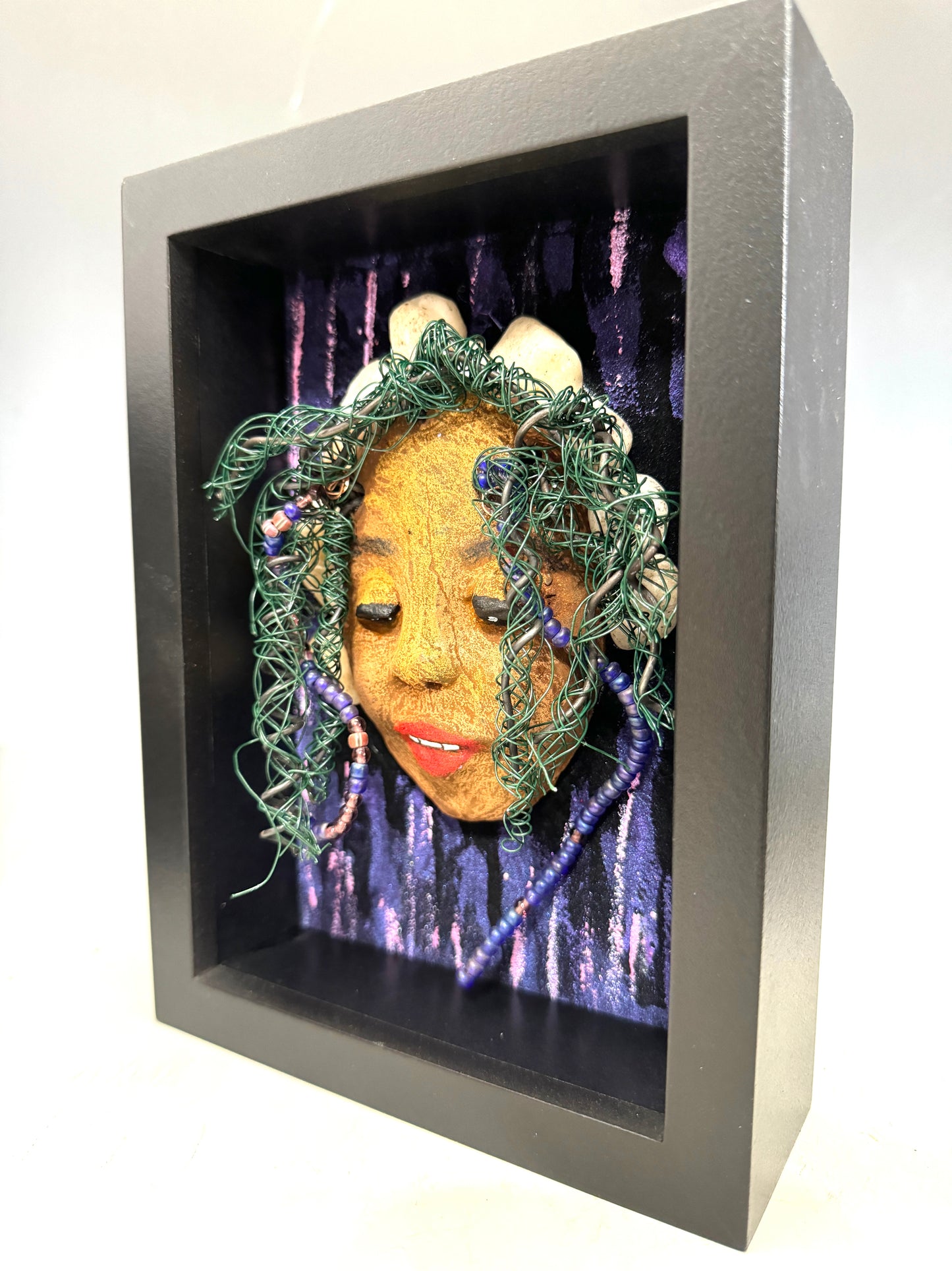 Presenting Shelby: A captivating mixed media sculpture made with a raku firing technique. Meticulously crafted features include a unique two-tone honey brown complexion, subtle Ruby red lips, and hair intricately woven with 16 and 24 gauge wire. This piece is mounted on a hand-painted canvas, featuring a beautifully landscaped background, and elegantly displayed in a glass covered 5" x 7" black shadowbox.