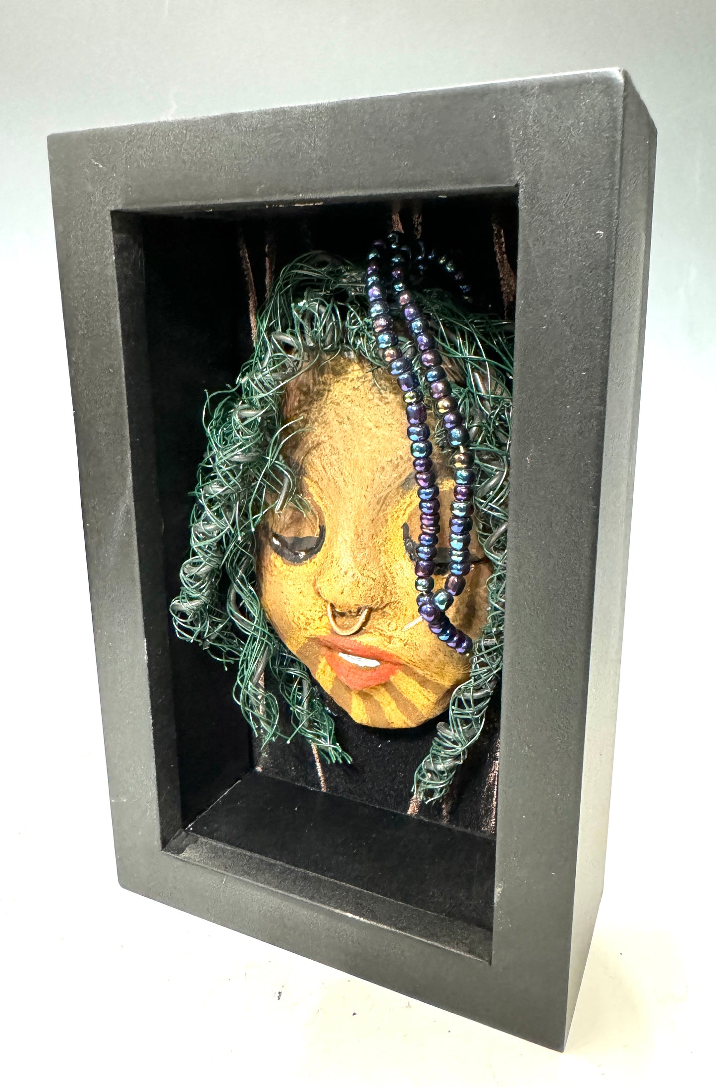 ntroducing Baraka: An alluring mixed media sculpture created using the raku firing method. Impeccably designed attributes include a distinctive dual-toned honey brown complexion, delicate Ruby red lips, and intricately woven hair using 16 and 24 gauge wire. Mounted on a hand-painted canvas with a stunning landscape backdrop, this piece is elegantly showcased in a 5" x 7" black shadowbox, protected by a glass cover.