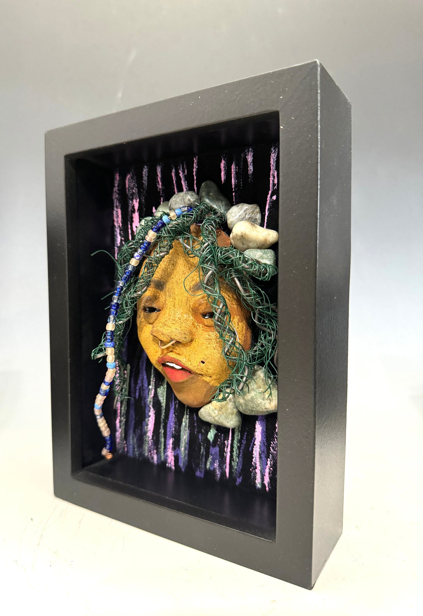 Introducing Beverly: A striking mixed media mask created using the raku firing method. The meticulously designed features showcase a beautiful honey brown complexion with subtle Ruby red lips and intricately woven hair made with 16 and 24 gauge wire. This exquisite piece is mounted on a hand-painted canvas, featuring a stunningly landscaped background, and elegantly presented in a glass covered 5" x 7" black shadowbox.