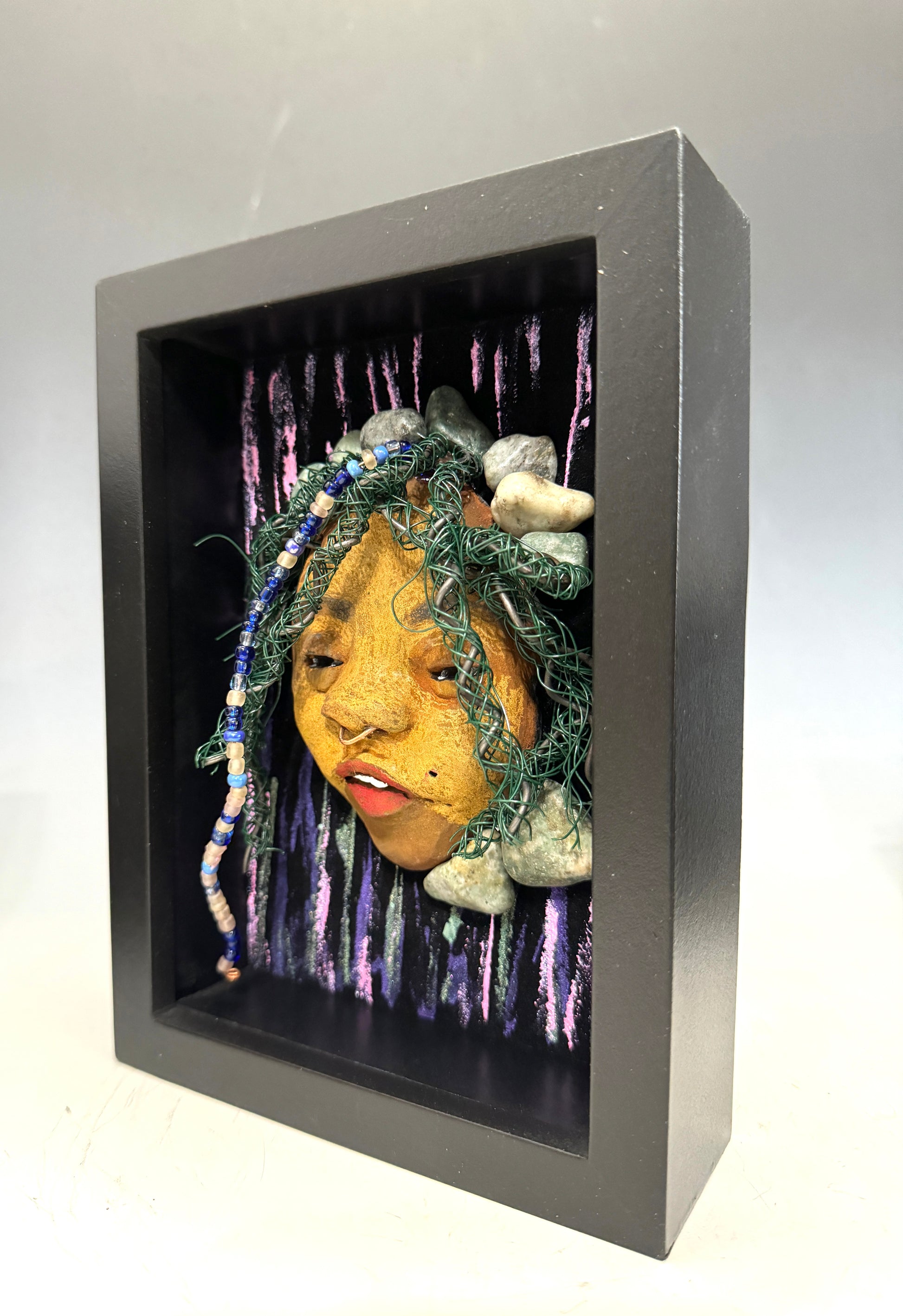 Introducing Beverly: A striking mixed media mask created using the raku firing method. The meticulously designed features showcase a beautiful honey brown complexion with subtle Ruby red lips and intricately woven hair made with 16 and 24 gauge wire. This exquisite piece is mounted on a hand-painted canvas, featuring a stunningly landscaped background, and elegantly presented in a glass covered 5" x 7" black shadowbox.