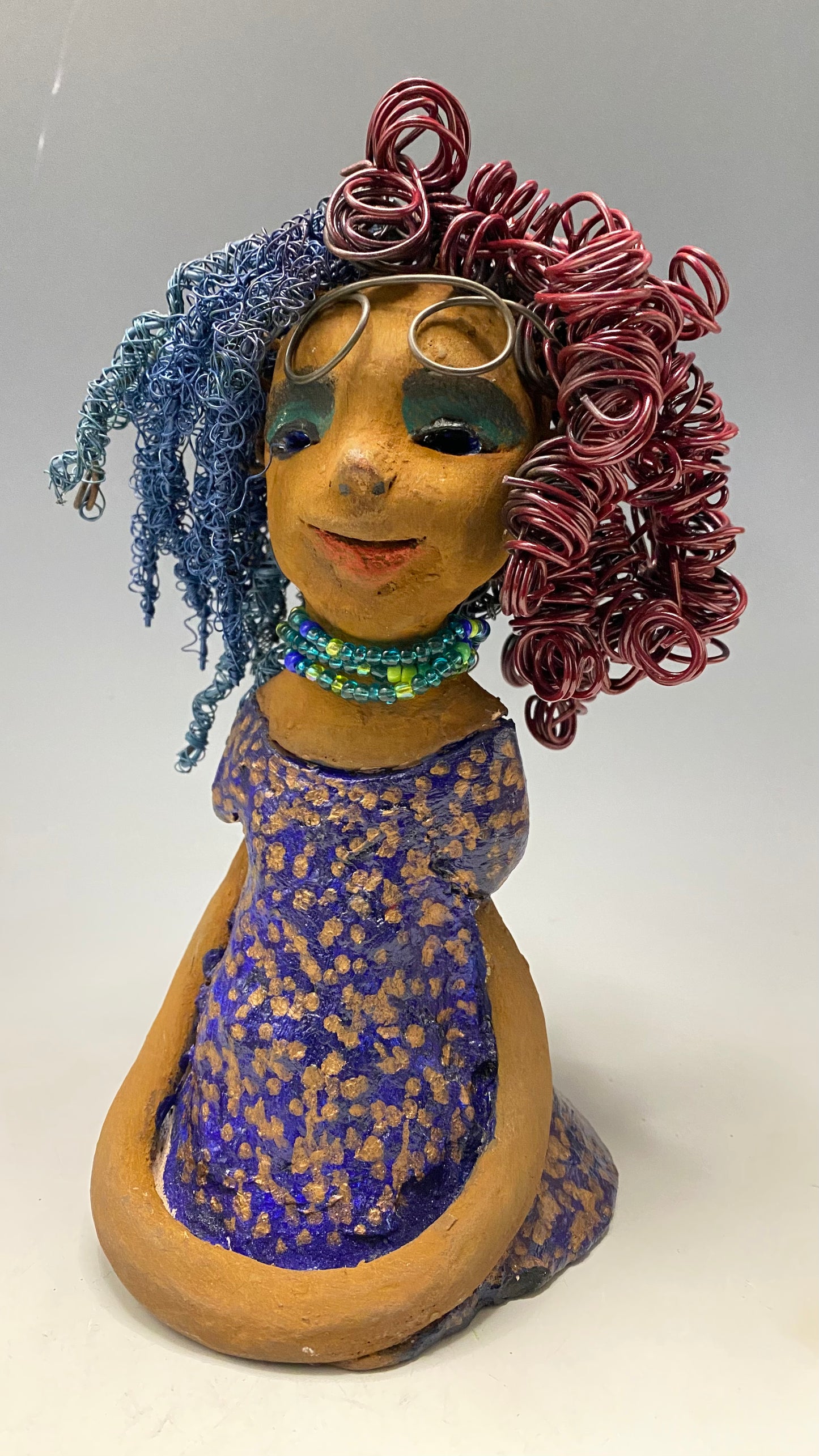 Cristy is 9" x 6" x 5.5", weighs 2.5 lbs, and has a honey brown complexion with reddish brown lips. Her long hair is adorned with red and blue color and 80 feet of 24 and 16 gauge twisted wire. She wears an antique glazed dress featuring a spotted blue and gold pattern. Cristy is completed by her eyeglasses which sit upon her forehead, inspiring hope and sparking conversations. Visit us for questions