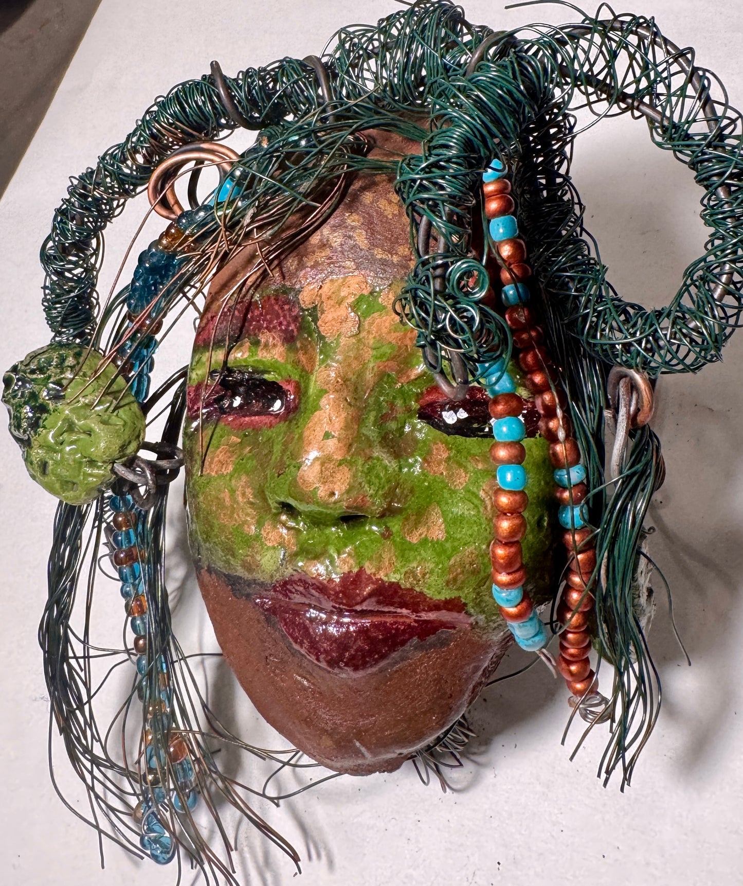 Taja is a Raku-fired mask measuring 4" x 5" x 2", featuring both 24 and 16 gauge wire to represent hair. This entrancing piece from the HerDew collection is the perfect gift for any holiday.