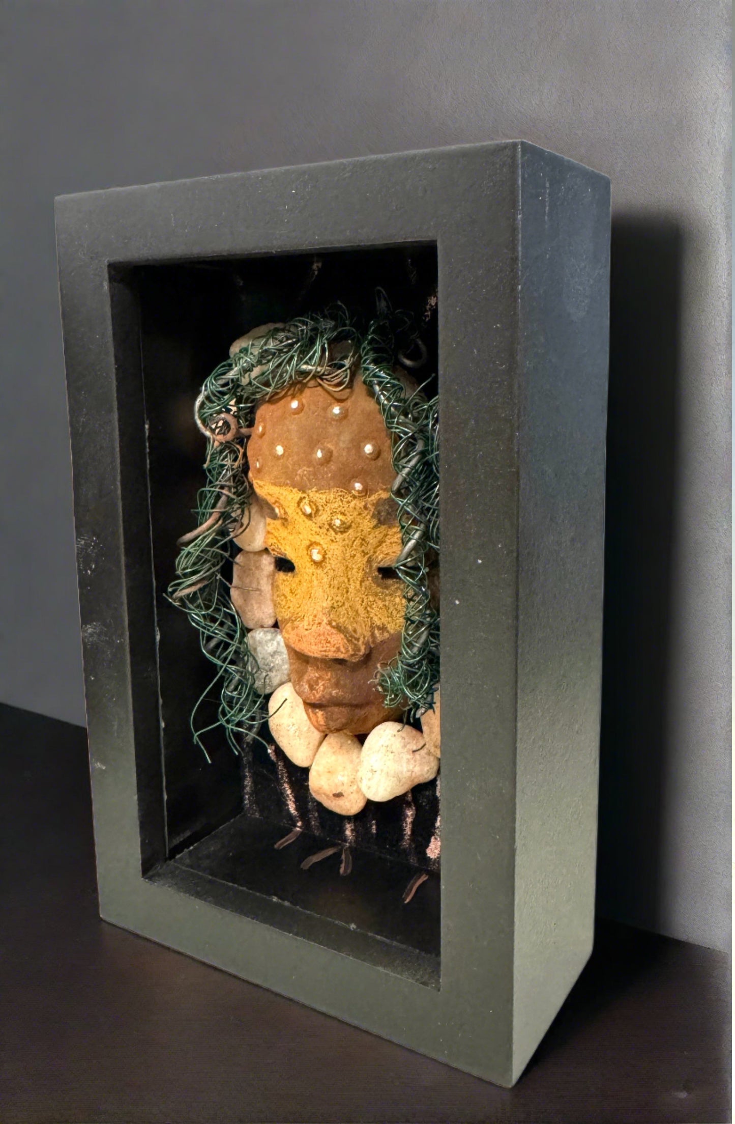 Presenting Craig: A captivating mixed media mask made with a raku firing technique. Meticulously crafted features include a unique two-tone honey brown complexion, subtle rust brown red lips, and hair intricately woven with 16 and 24 gauge wire. This piece is mounted on a hand-painted canvas, featuring a beautifully landscaped background, and elegantly displayed in a glass covered 4" x 6" black shadowbox.