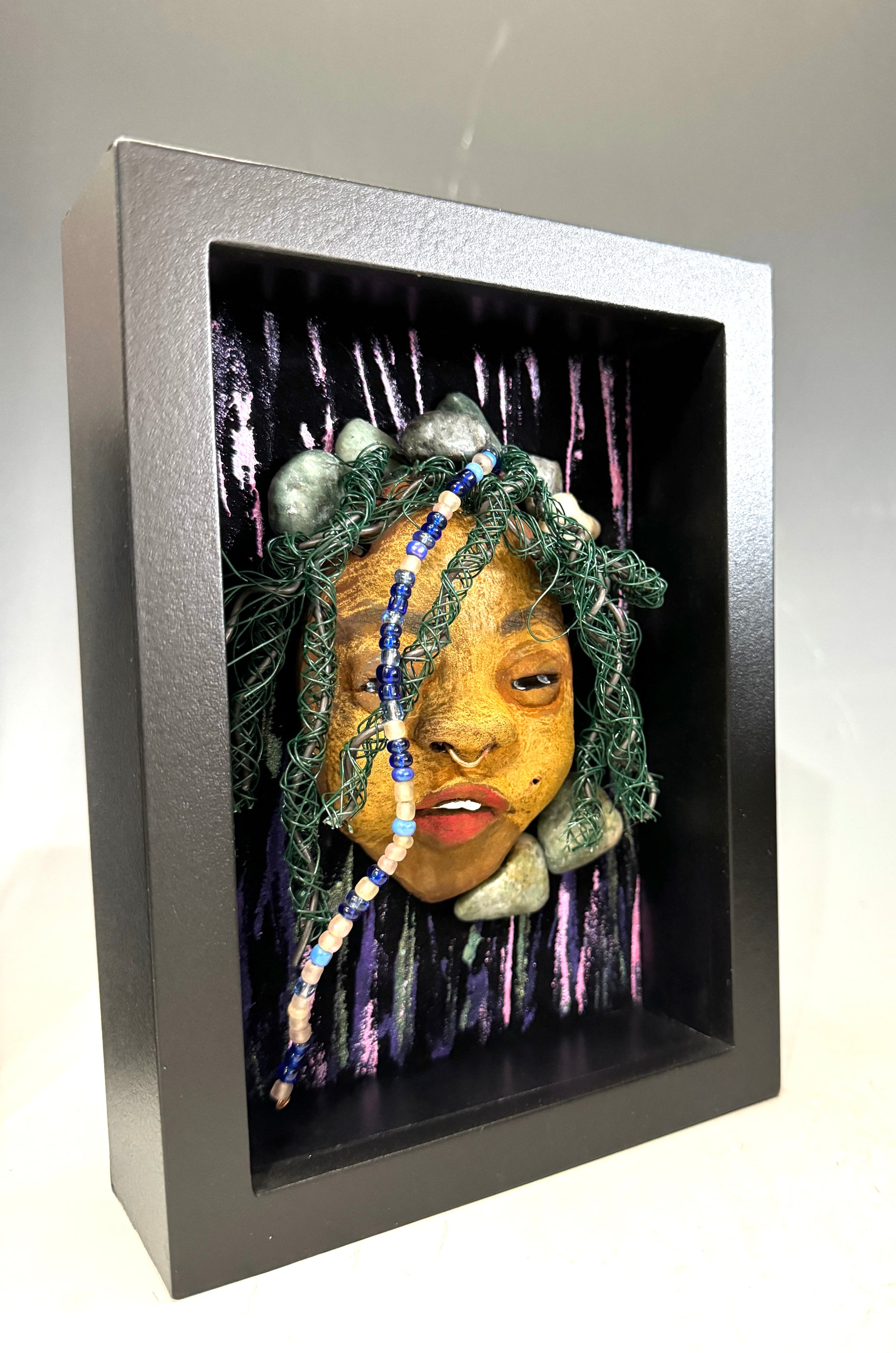 Introducing Beverly: A striking mixed media mask created using the raku firing method. The meticulously designed features showcase a beautiful honey brown complexion with subtle Ruby red lips and intricately woven hair made with 16 and 24 gauge wire. This exquisite piece is mounted on a hand-painted canvas, featuring a stunningly landscaped background, and elegantly presented in a glass covered 5" x 7" black shadowbox.