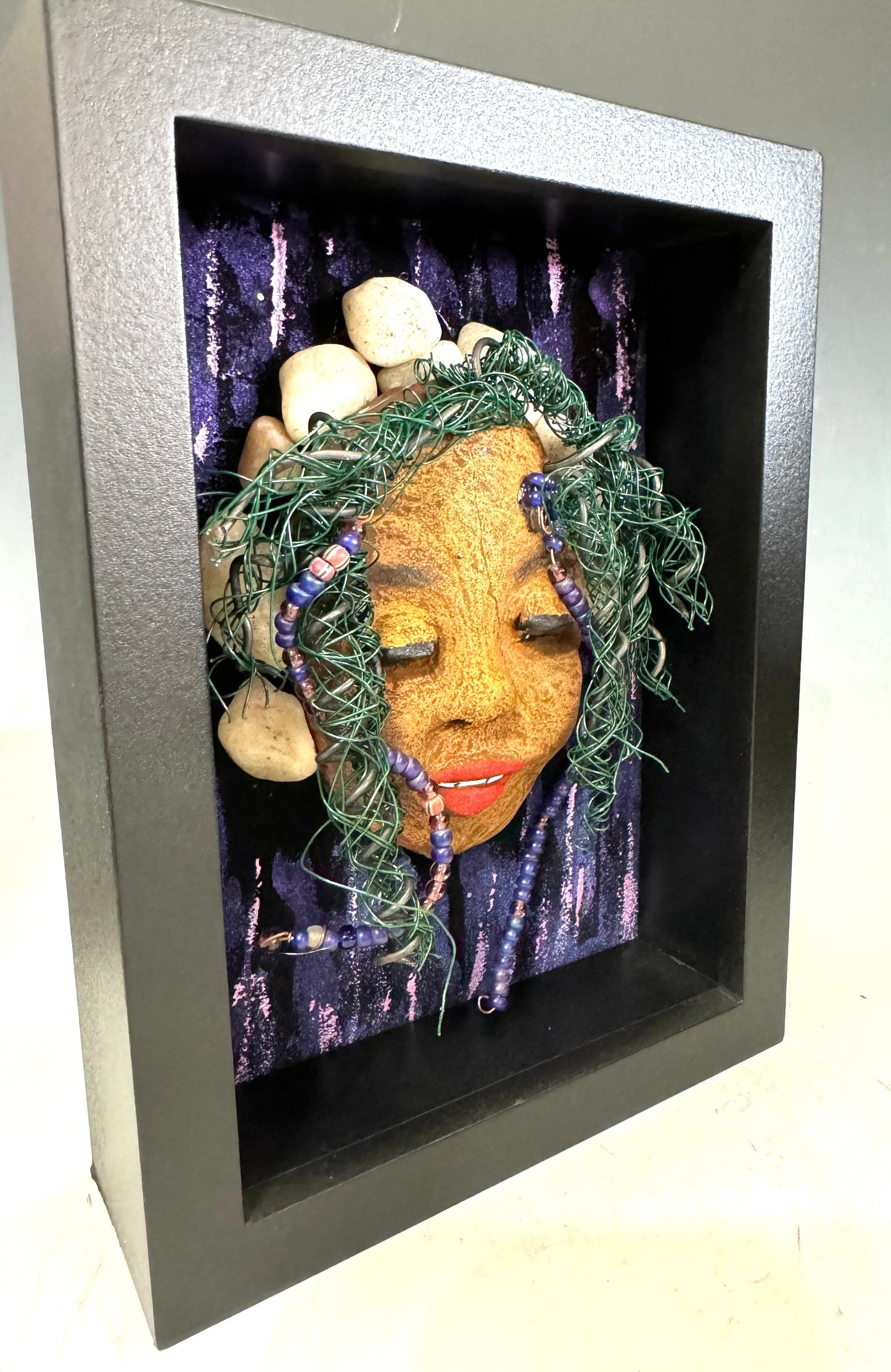 Presenting Shelby: A captivating mixed media sculpture made with a raku firing technique. Meticulously crafted features include a unique two-tone honey brown complexion, subtle Ruby red lips, and hair intricately woven with 16 and 24 gauge wire. This piece is mounted on a hand-painted canvas, featuring a beautifully landscaped background, and elegantly displayed in a glass covered 5" x 7" black shadowbox.