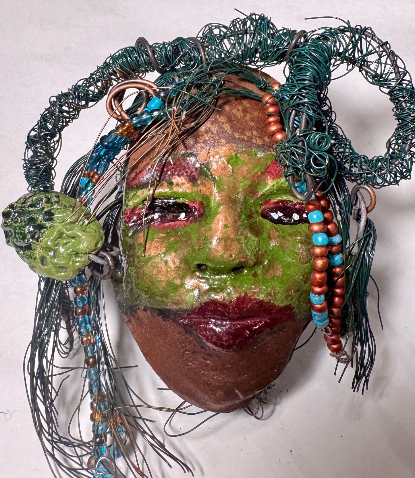 Taja is a Raku-fired mask measuring 4" x 5" x 2", featuring both 24 and 16 gauge wire to represent hair. This entrancing piece from the HerDew collection is the perfect gift for any holiday.