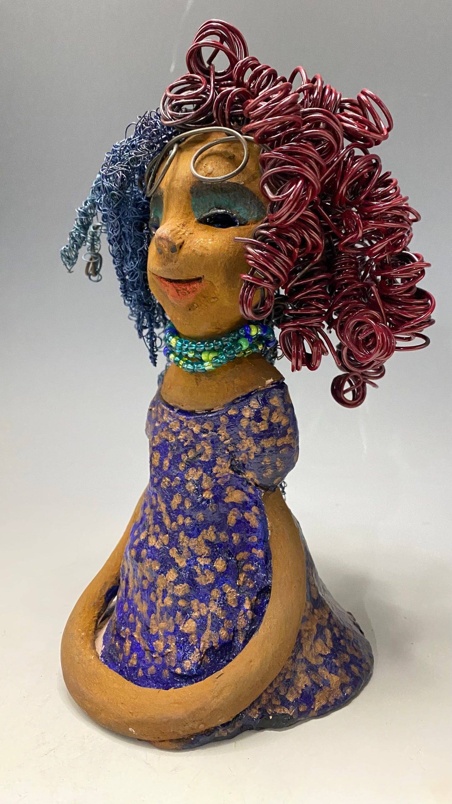 Cristy is 9" x 6" x 5.5", weighs 2.5 lbs, and has a honey brown complexion with reddish brown lips. Her long hair is adorned with red and blue color and 80 feet of 24 and 16 gauge twisted wire. She wears an antique glazed dress featuring a spotted blue and gold pattern. Cristy is completed by her eyeglasses which sit upon her forehead, inspiring hope and sparking conversations. Visit us for questions