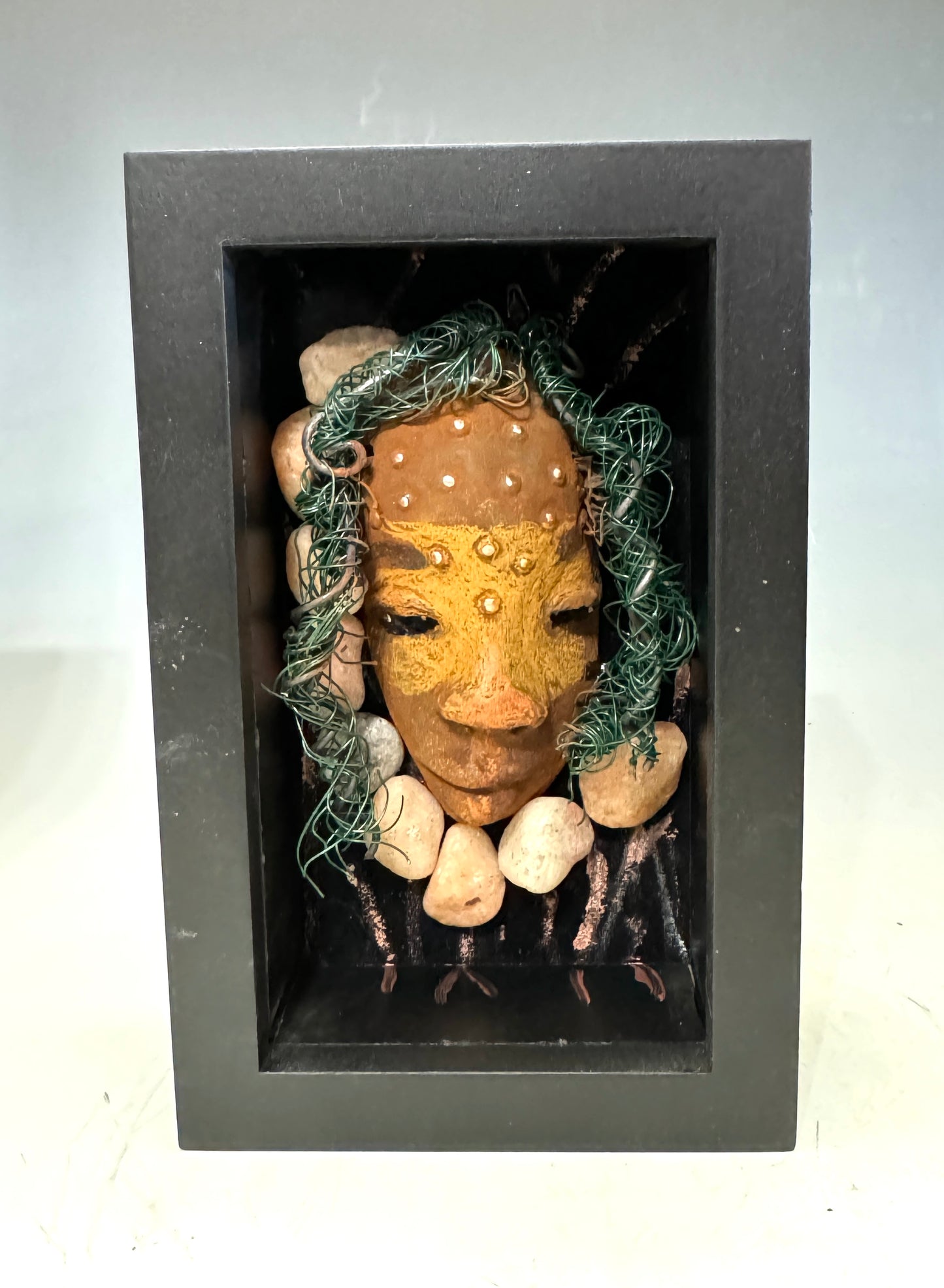 Presenting Craig: A captivating mixed media mask made with a raku firing technique. Meticulously crafted features include a unique two-tone honey brown complexion, subtle rust brown red lips, and hair intricately woven with 16 and 24 gauge wire. This piece is mounted on a hand-painted canvas, featuring a beautifully landscaped background, and elegantly displayed in a glass covered 4" x 6" black shadowbox.