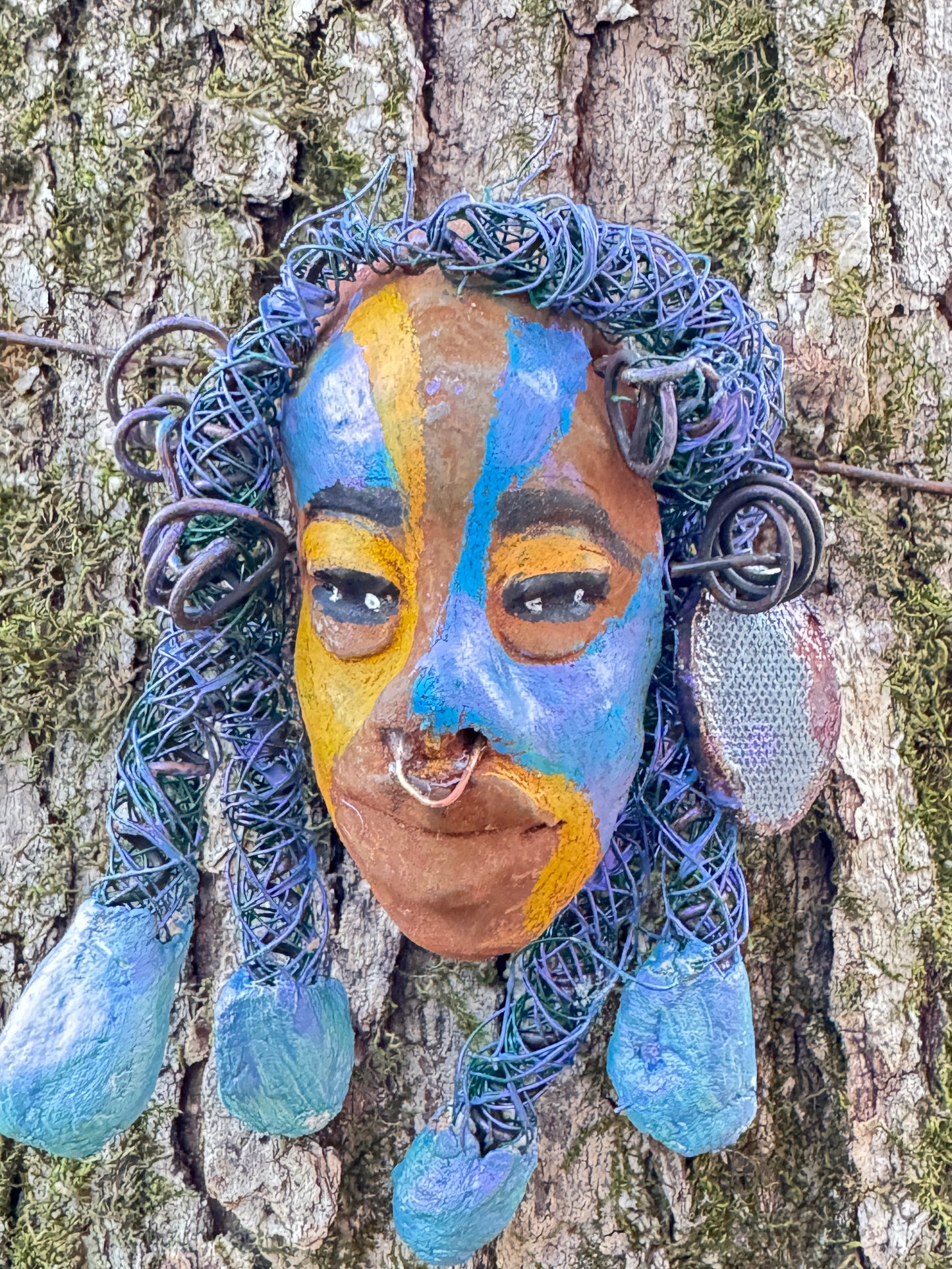 Esi is a lightweight 4 x 6" mixed media raku fire mask that combines a striking design with 24 and 16 gauge wire, 5 intricate blue tribal beads, and over 15 feet of wire. It can be proudly displayed against a solid black or white background, or for an even more impressive effect, consider a backdrop featuring lush, mossy trees.