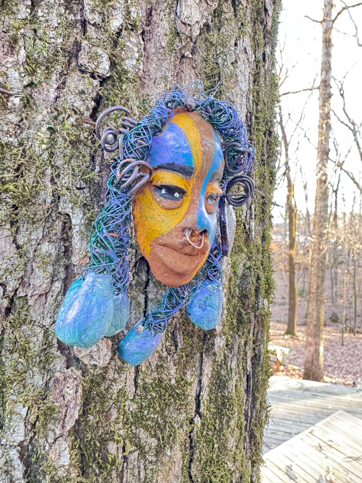 Esi is a lightweight 4 x 6" mixed media raku fire mask that combines a striking design with 24 and 16 gauge wire, 5 intricate blue tribal beads, and over 15 feet of wire. It can be proudly displayed against a solid black or white background, or for an even more impressive effect, consider a backdrop featuring lush, mossy trees.