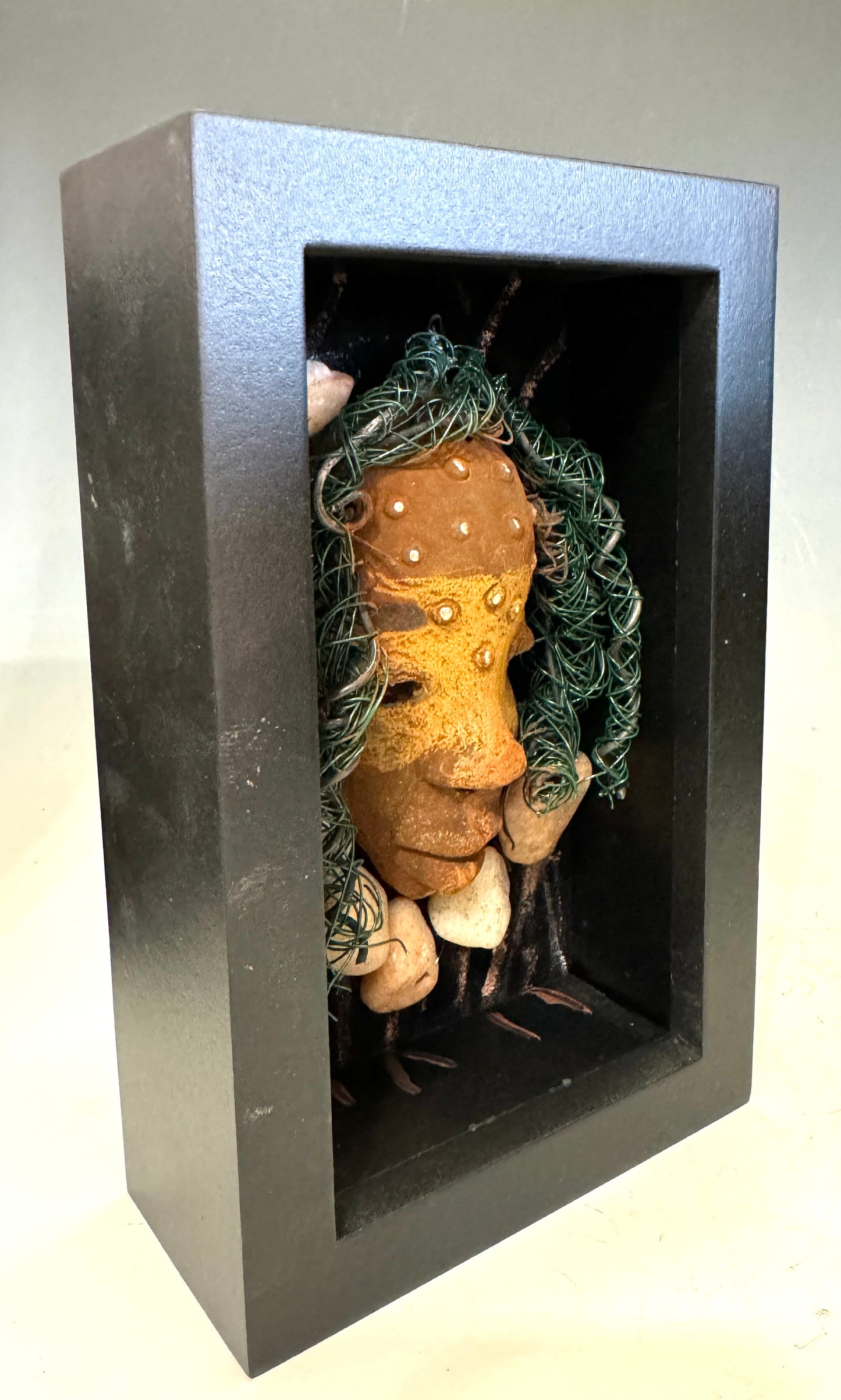 Presenting Craig: A captivating mixed media mask made with a raku firing technique. Meticulously crafted features include a unique two-tone honey brown complexion, subtle rust brown red lips, and hair intricately woven with 16 and 24 gauge wire. This piece is mounted on a hand-painted canvas, featuring a beautifully landscaped background, and elegantly displayed in a glass covered 4" x 6" black shadowbox.