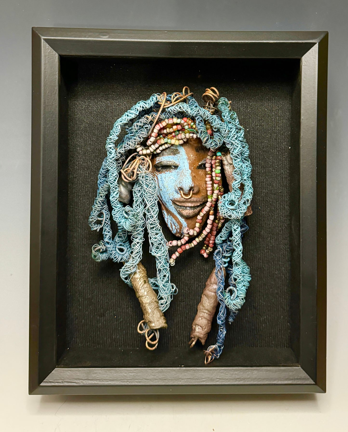 Zola is a mesmerizing raku mask with a captivating tribal face. She measures approximately 5 x 9" and weighs 7 oz. The mask is further decorated with blue hair and a copper nose ring.