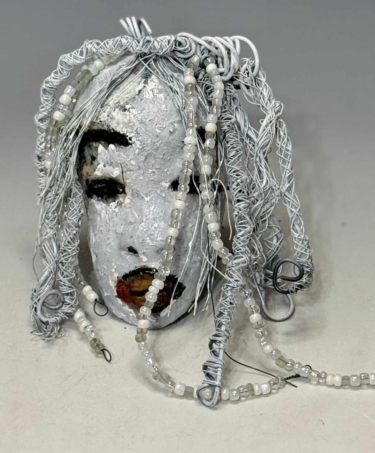 Olivia is a 4 x 7” 5.5 oz raku-fired mask featuring an off-white complexion. 16 gauge wire measuring over 15 feet is used as her hair, accessorized with small decorative beads. Her portrait was taken in an outdoor setting featuring a tree, making her an ideal gift for those who enjoy outdoor art or want to adorn the inside of their home.