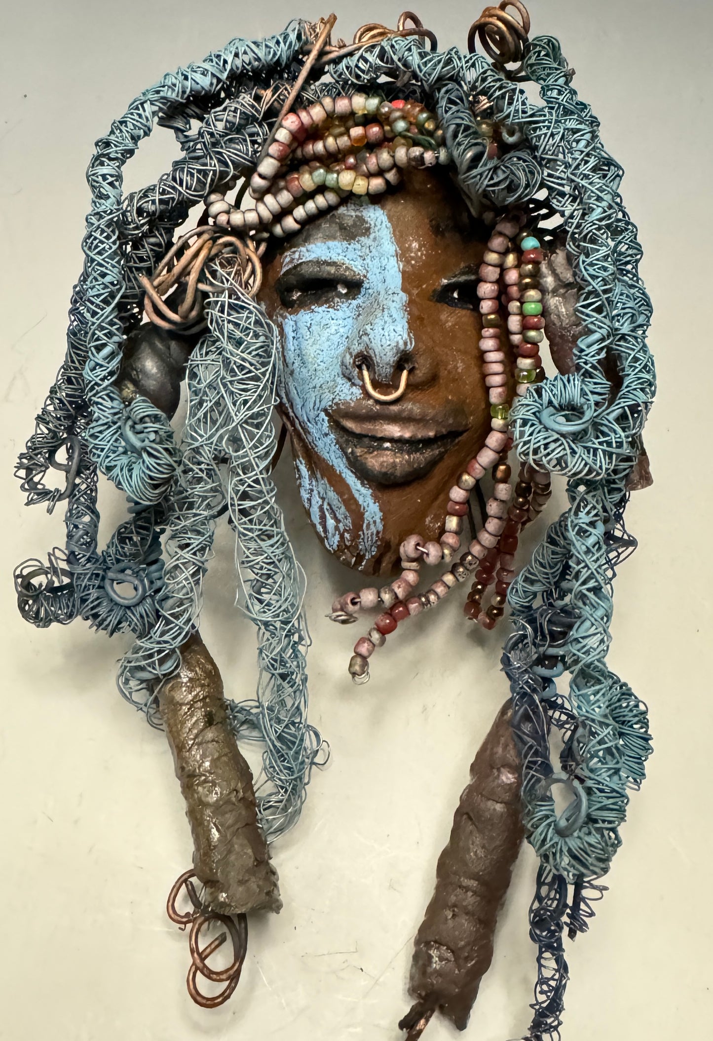 Zola is a mesmerizing raku mask with a captivating tribal face. She measures approximately 5 x 9" and weighs 7 oz. The mask is further decorated with blue hair and a copper nose ring.