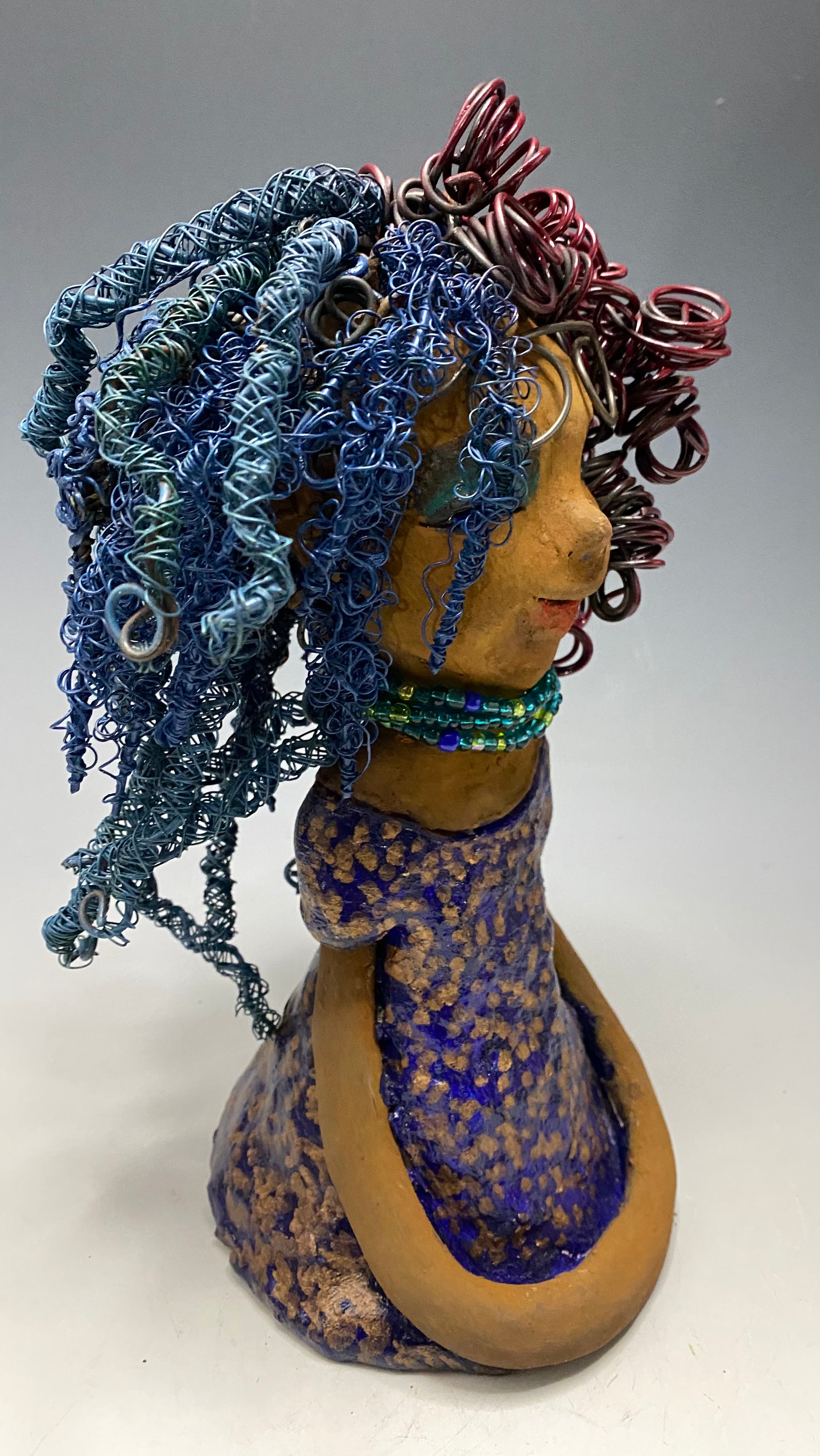 Cristy is 9" x 6" x 5.5", weighs 2.5 lbs, and has a honey brown complexion with reddish brown lips. Her long hair is adorned with red and blue color and 80 feet of 24 and 16 gauge twisted wire. She wears an antique glazed dress featuring a spotted blue and gold pattern. Cristy is completed by her eyeglasses which sit upon her forehead, inspiring hope and sparking conversations. Visit us for questions