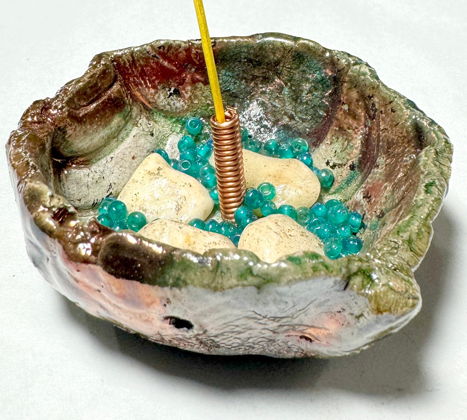 This 1" x 3" x 3" incense holder has been expertly crafted with earthy off-white stones and aqua blue beads and coated in a copper high gloss Raku glaze.