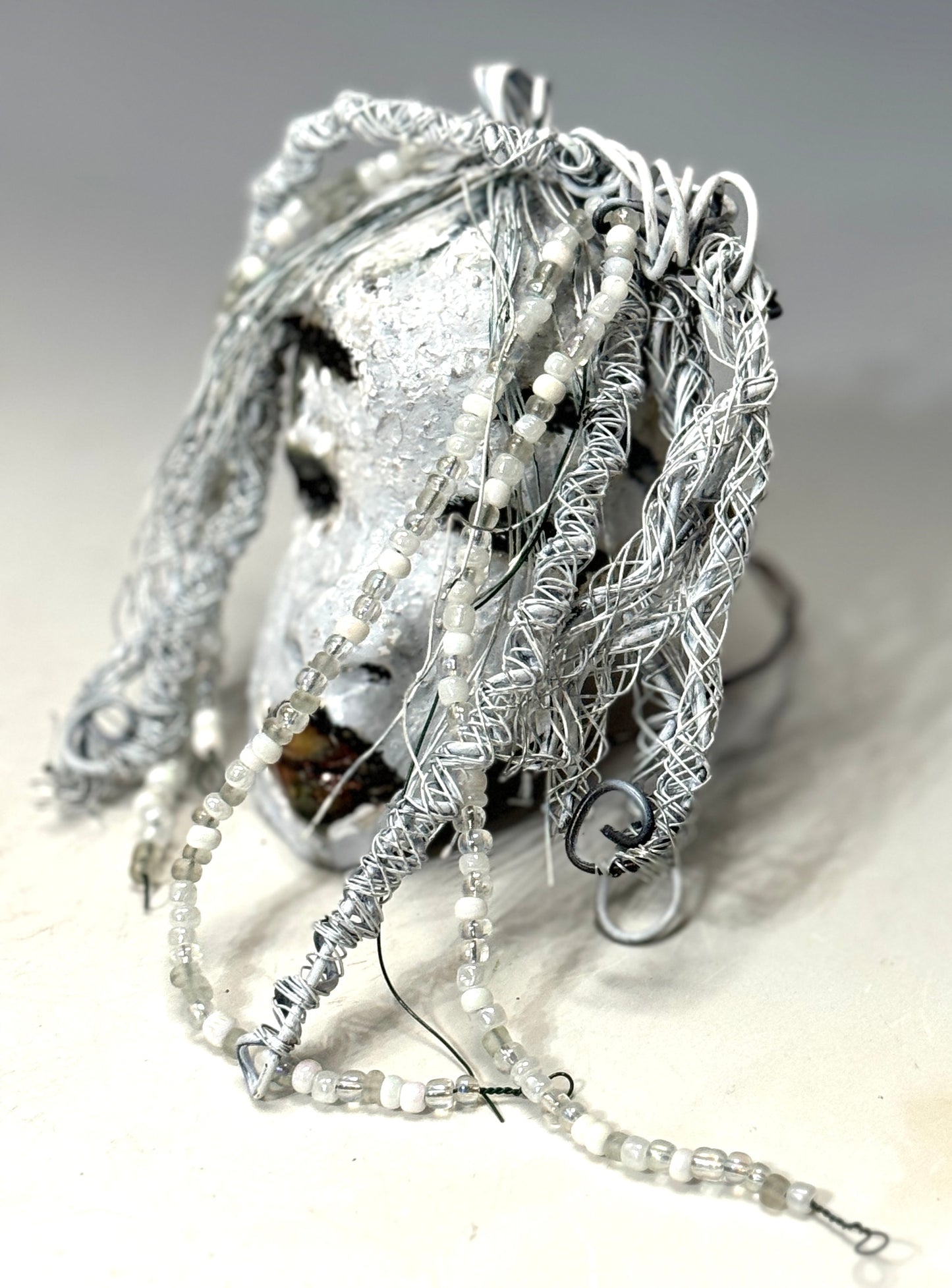 Olivia is a 4 x 7” 5.5 oz raku-fired mask featuring an off-white complexion. 16 gauge wire measuring over 15 feet is used as her hair, accessorized with small decorative beads. Her portrait was taken in an outdoor setting featuring a tree, making her an ideal gift for those who enjoy outdoor art or want to adorn the inside of their home.