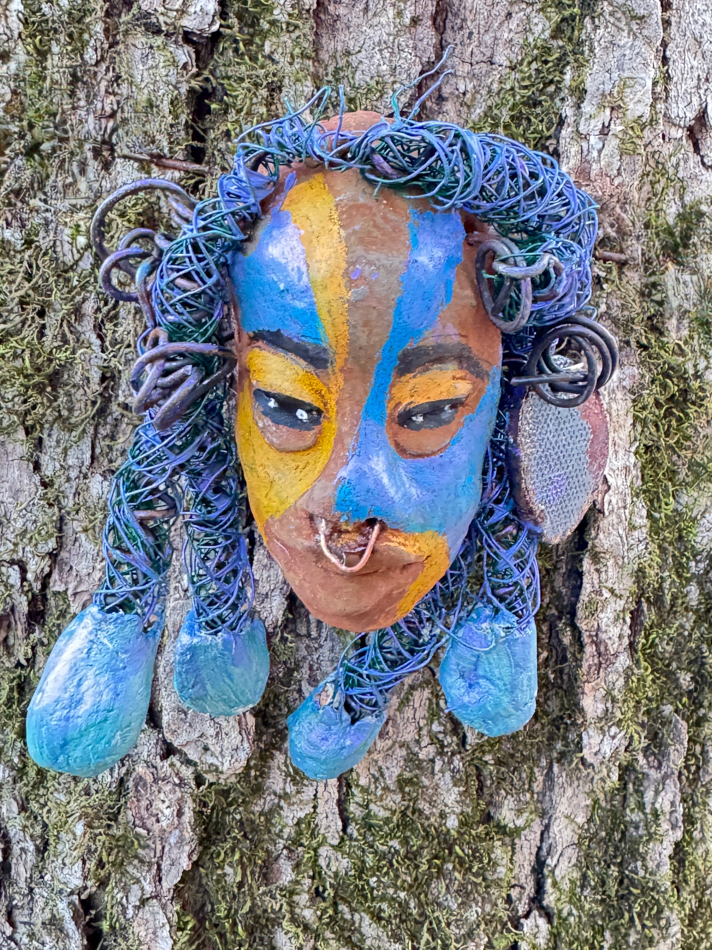 Esi is a lightweight 4 x 6" mixed media raku fire mask that combines a striking design with 24 and 16 gauge wire, 5 intricate blue tribal beads, and over 15 feet of wire. It can be proudly displayed against a solid black or white background, or for an even more impressive effect, consider a backdrop featuring lush, mossy trees.