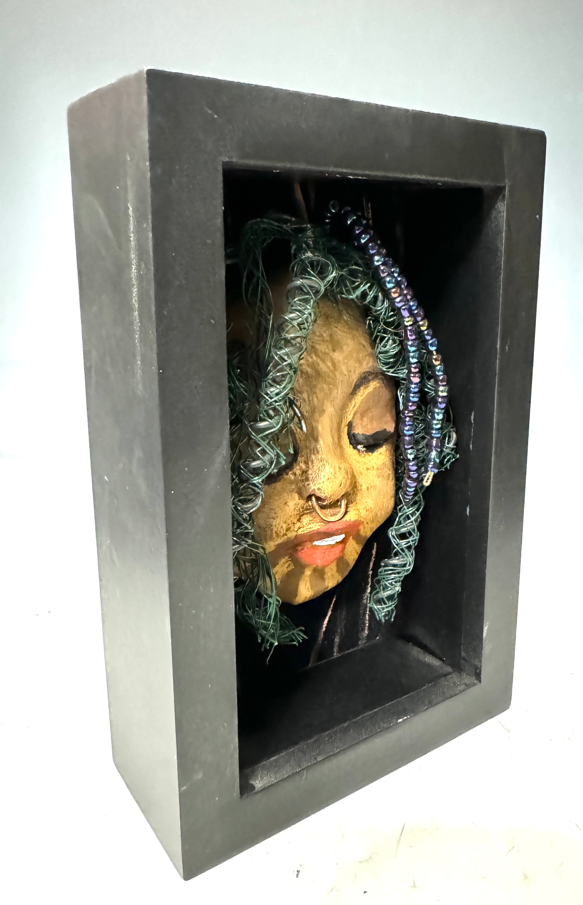 ntroducing Baraka: An alluring mixed media sculpture created using the raku firing method. Impeccably designed attributes include a distinctive dual-toned honey brown complexion, delicate Ruby red lips, and intricately woven hair using 16 and 24 gauge wire. Mounted on a hand-painted canvas with a stunning landscape backdrop, this piece is elegantly showcased in a 5" x 7" black shadowbox, protected by a glass cover.