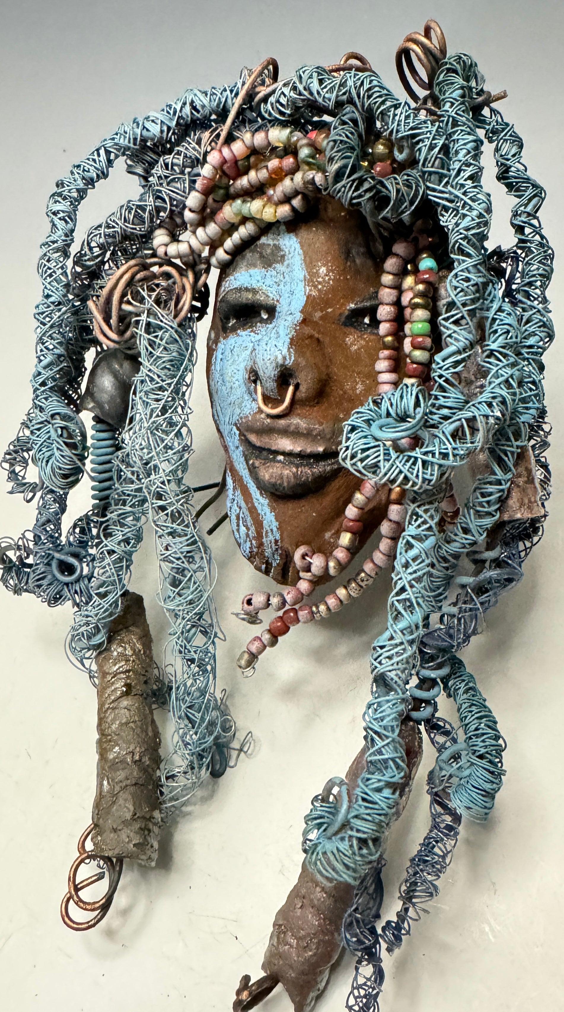 Zola is a mesmerizing raku mask with a captivating tribal face. She measures approximately 5 x 9" and weighs 7 oz. The mask is further decorated with blue hair and a copper nose ring.