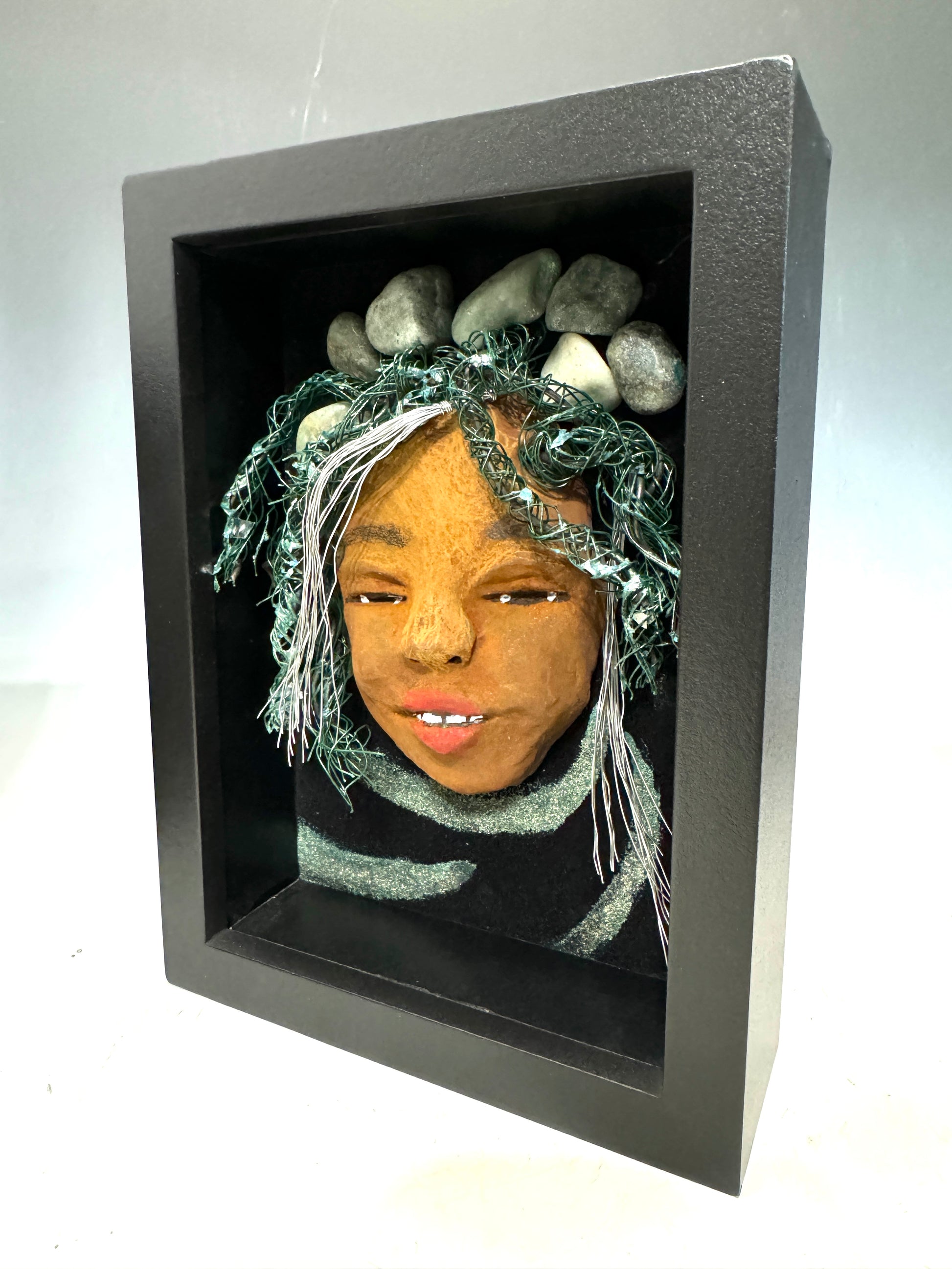 Introducing Brandie, a mixed media sculptured raku fired mask. Her features include a two-tone honey brown complexion, Ruby red lips, and hair intricately intertwined with 16 and 24 gauge wire. She is mounted on a hand-painted canvas, accented by an earthy landscape background, and enclosed in a 5“ x 7“ black shadowbox.