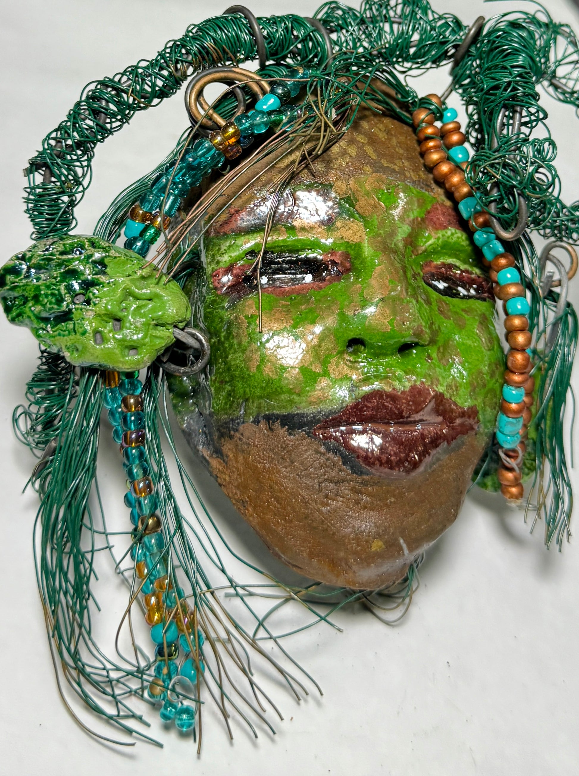Taja is a Raku-fired mask measuring 4" x 5" x 2", featuring both 24 and 16 gauge wire to represent hair. This entrancing piece from the HerDew collection is the perfect gift for any holiday.